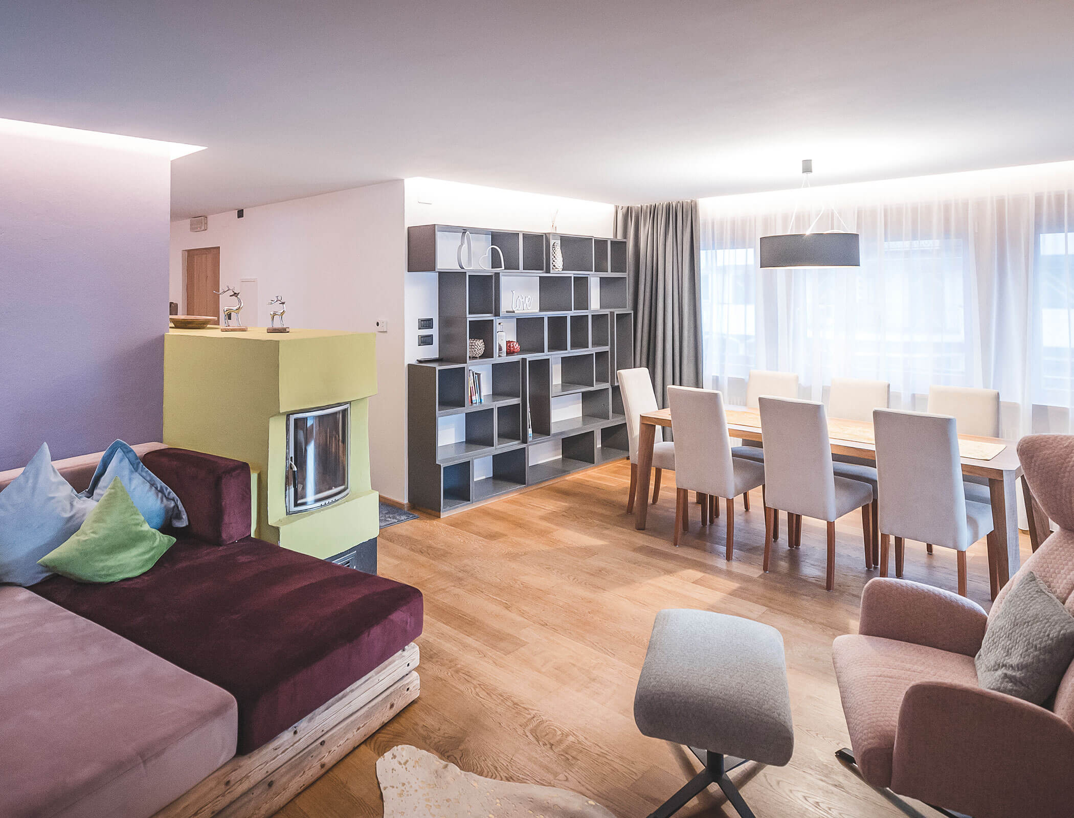 Modernes Wohn- und Esszimmer mit Holzböden, Möbeln in neutralen Farbtönen, offenen Regalen und großen Fenstern mit Vorhängen. - Hotel Fameli