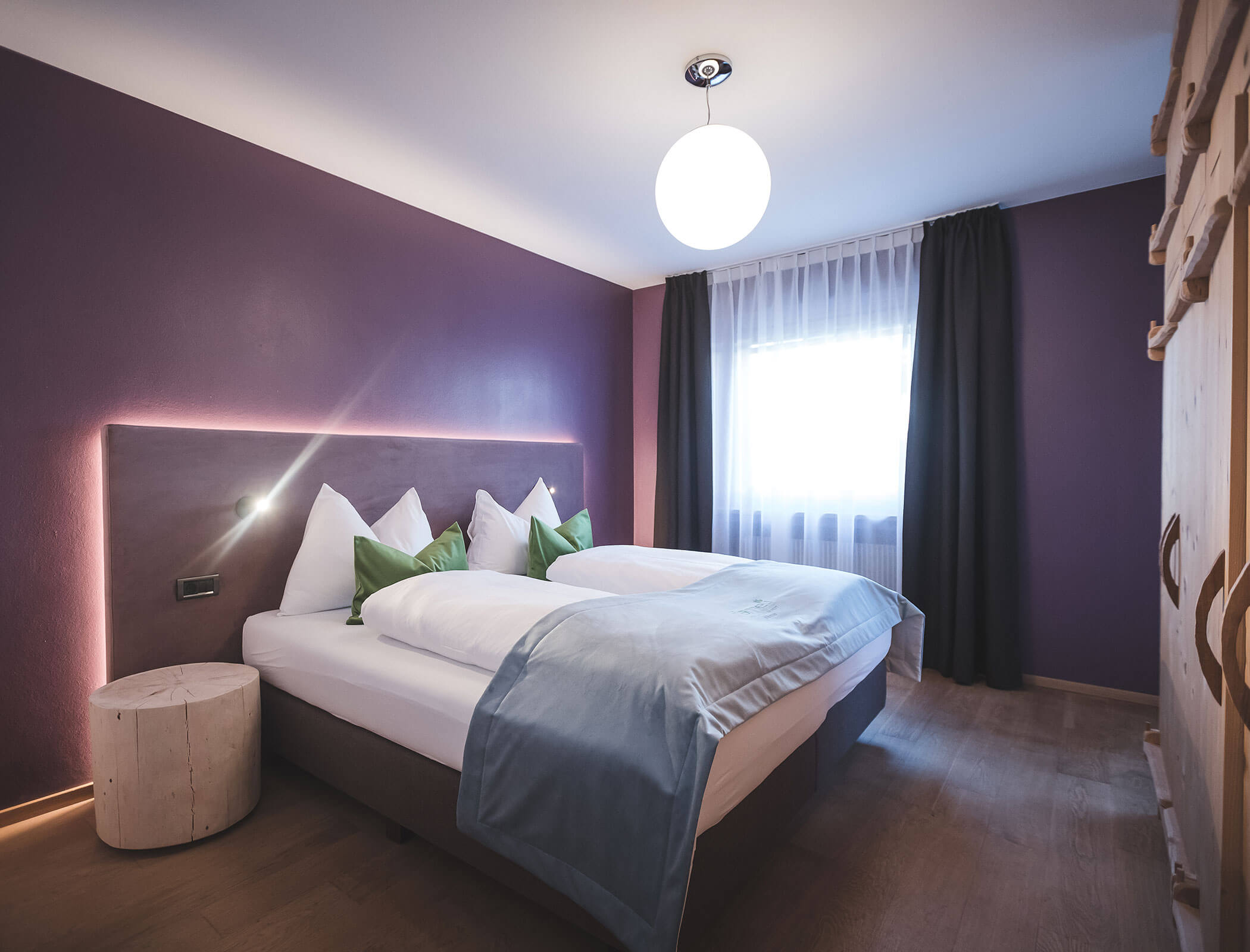 Modernes Schlafzimmer mit lila Wänden, einem Doppelbett, grünen Kissen und einem großen Fenster mit Vorhängen. - Hotel Fameli