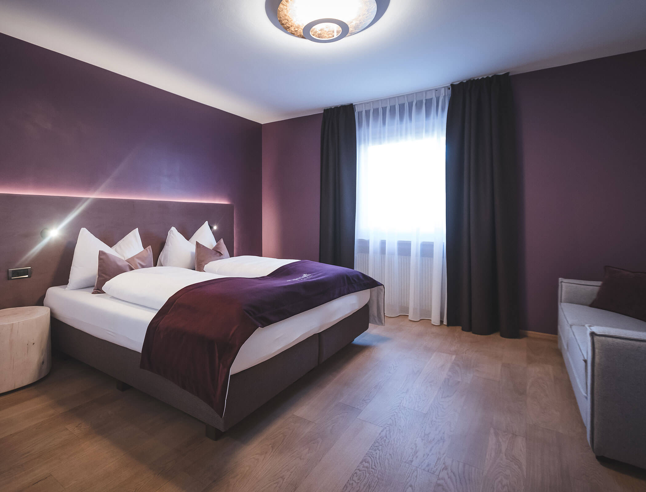 Ein modernes Schlafzimmer mit lila Wänden, einem Doppelbett, weißen Vorhängen und einem grauen Sofa auf einem Holzboden. - Hotel Fameli
