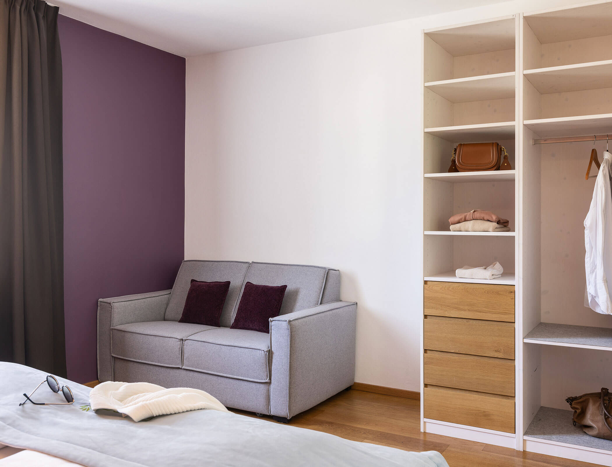 Graues Sofa mit violetten Kissen neben einem offenen Schrank und Holzkommoden in einem modernen Schlafzimmer. - Hotel Fameli