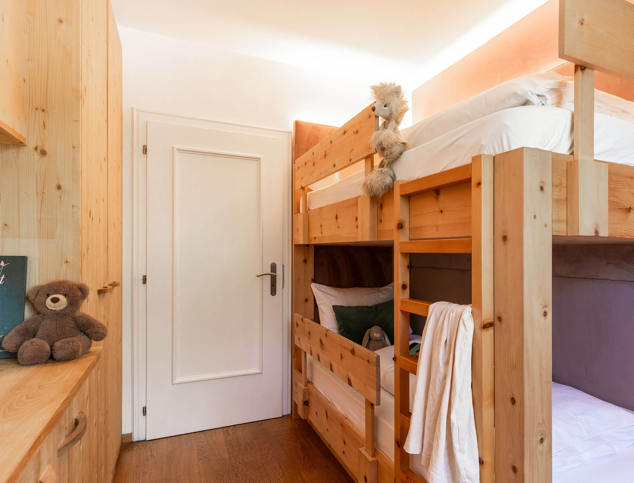 Ein kleines Zimmer aus Holz mit Etagenbetten, weißer Bettwäsche, Stofftieren und einer geschlossenen weißen Tür. - Hotel Fameli