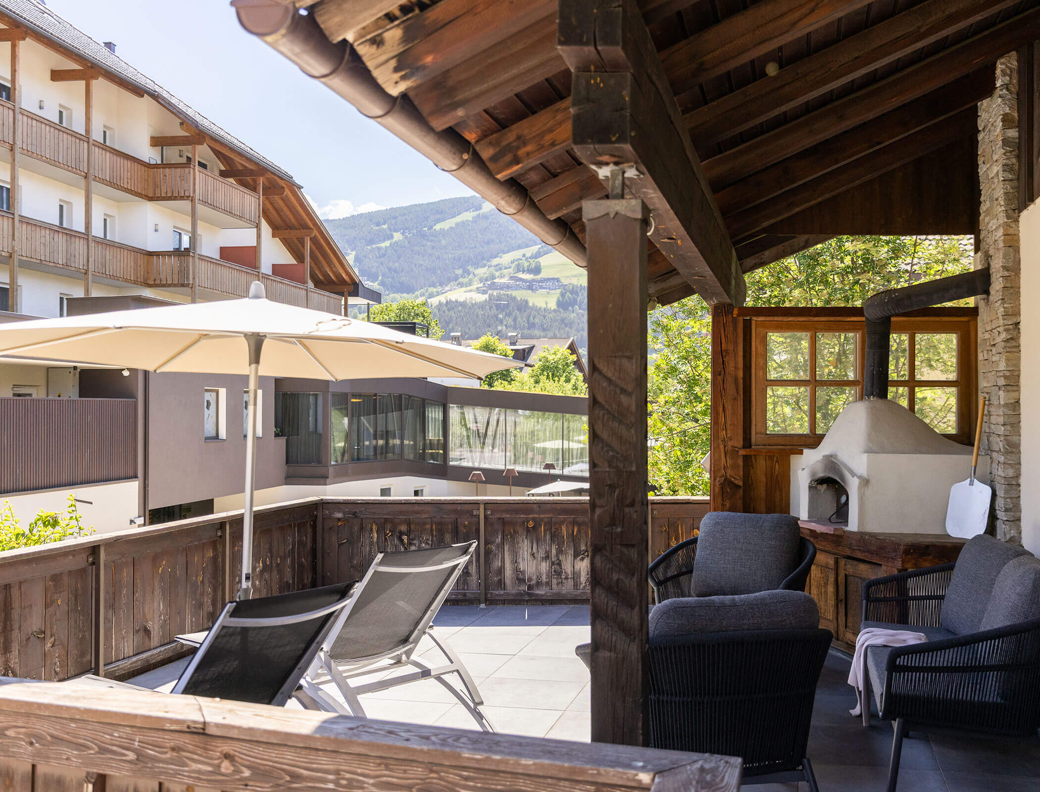 Terrasse mit Bergblick, Liegestühlen, einem Sonnenschirm und einem Außenofen unter einem Holzdach. - Hotel Fameli