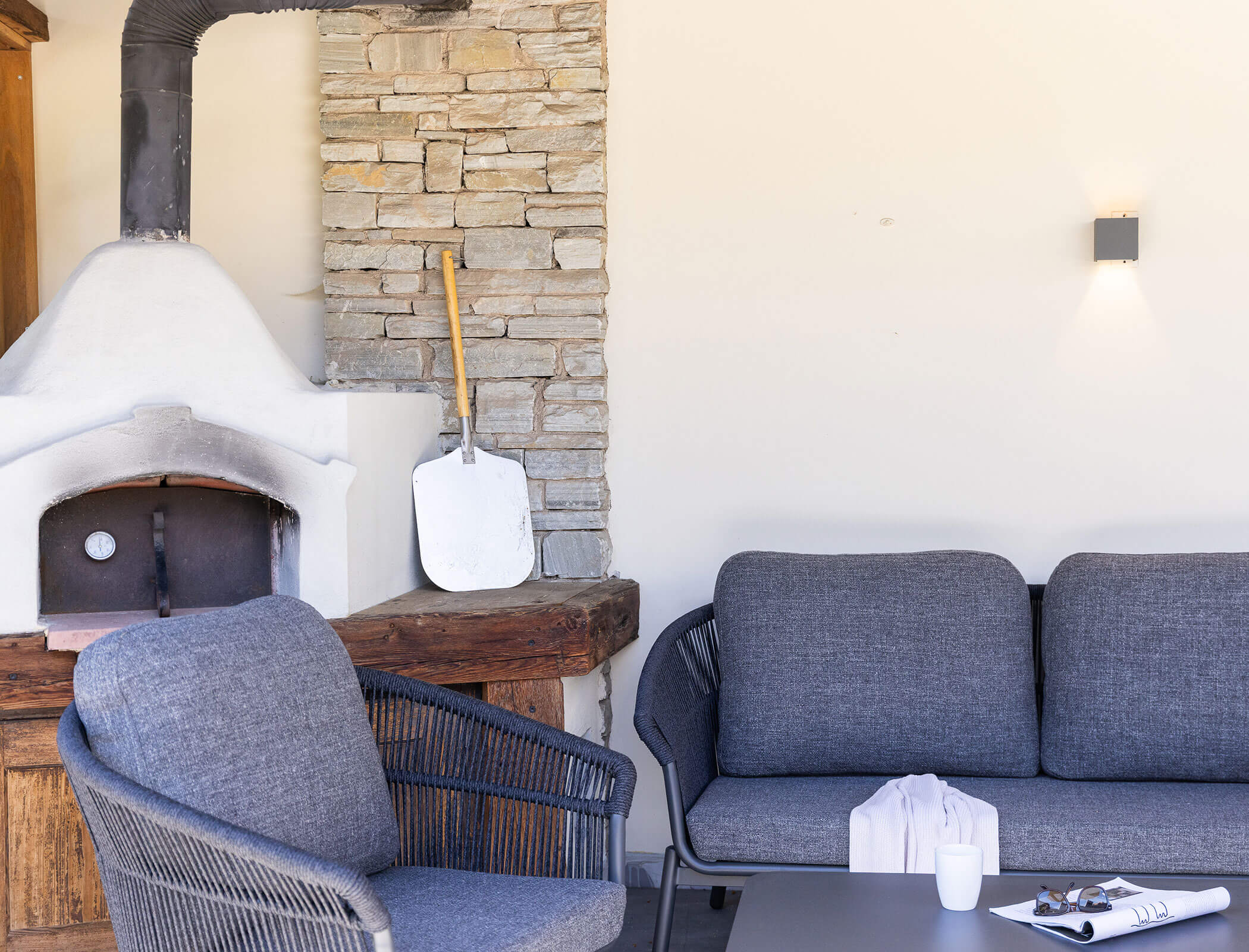 Graue Gartenstühle und ein Sofa neben einem Pizzaofen aus Stein, auf dem Tisch liegen eine Pizzaschaufel und eine Tasse. - Hotel Fameli