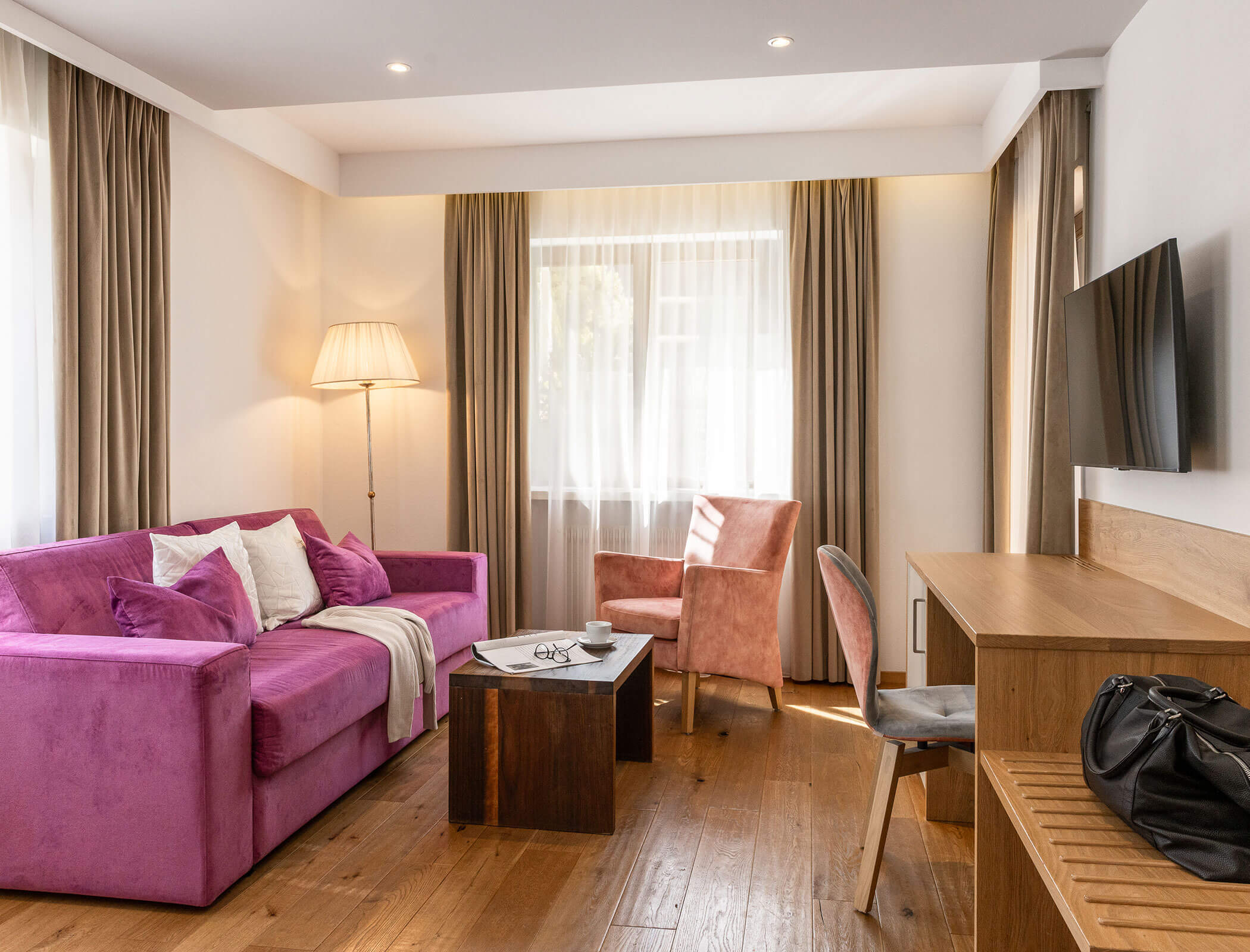 Bright living room with a pink sofa, armchair, desk, TV, and large windows with beige curtains. - Hotel Fameli