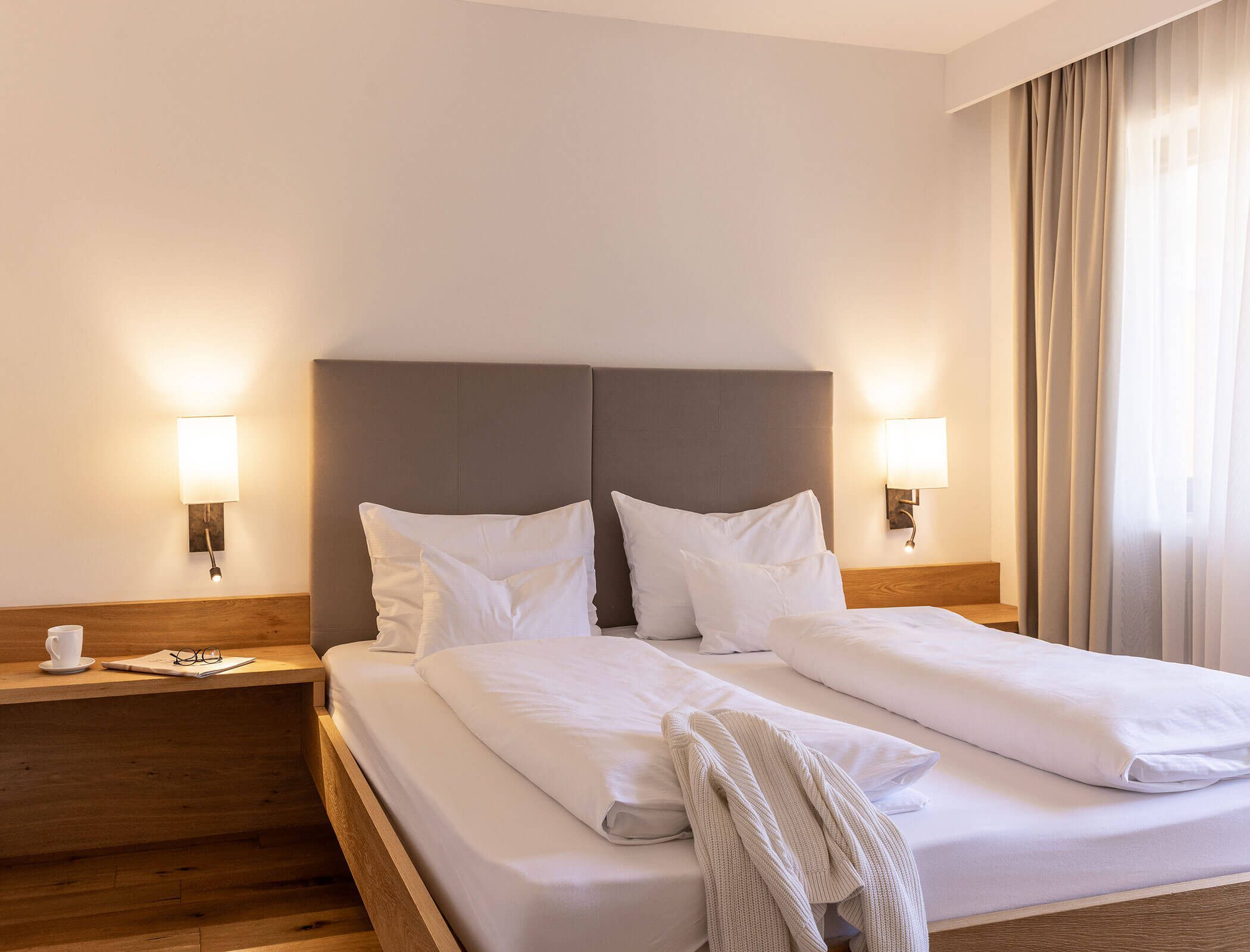 Hotel room with a double bed, two lamps, a robe, glasses, and a cup on a nightstand near a window. - Hotel Fameli