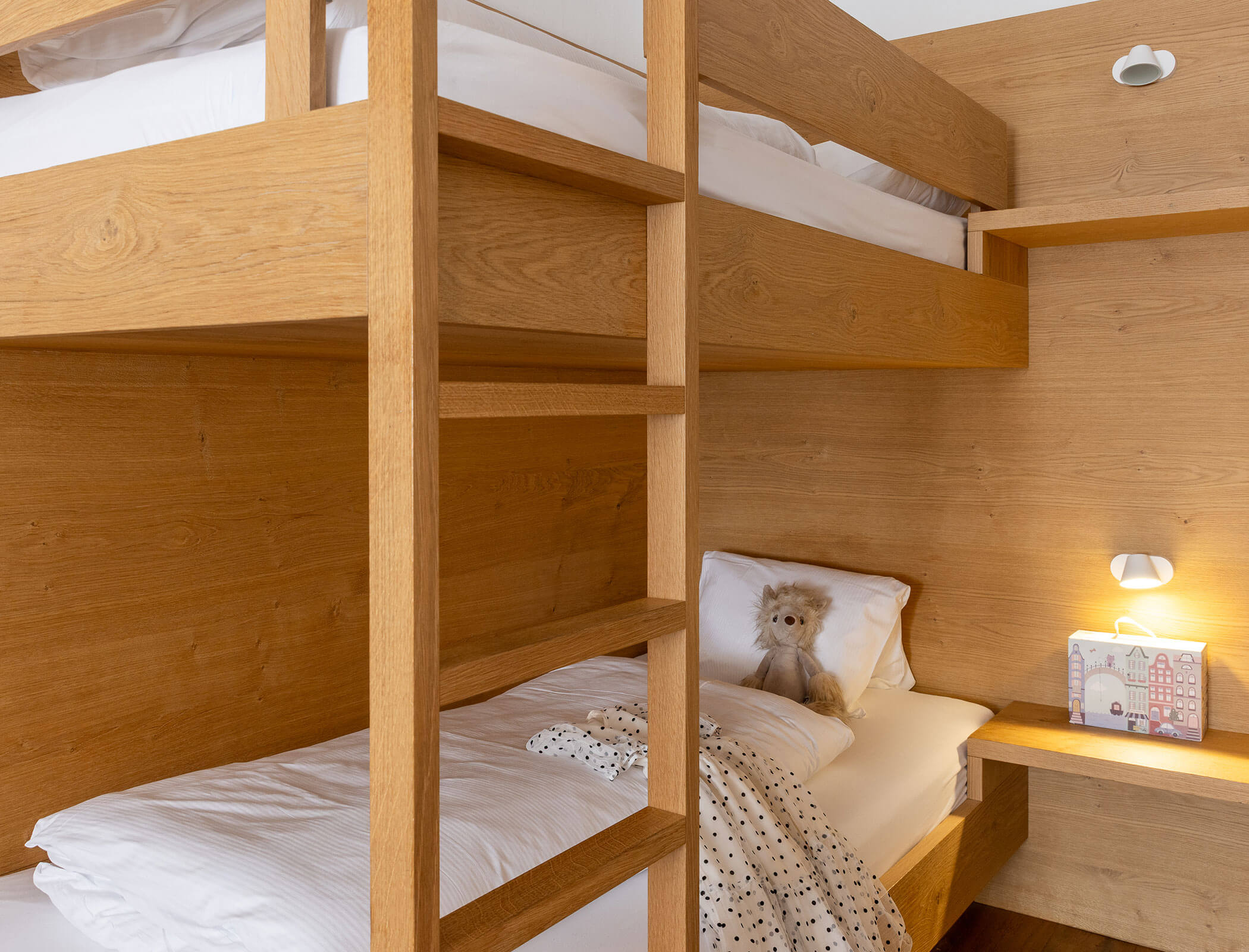 Wooden bunk beds with white bedding; a teddy bear sits on the lower bed next to a small lit desk. - Hotel Fameli