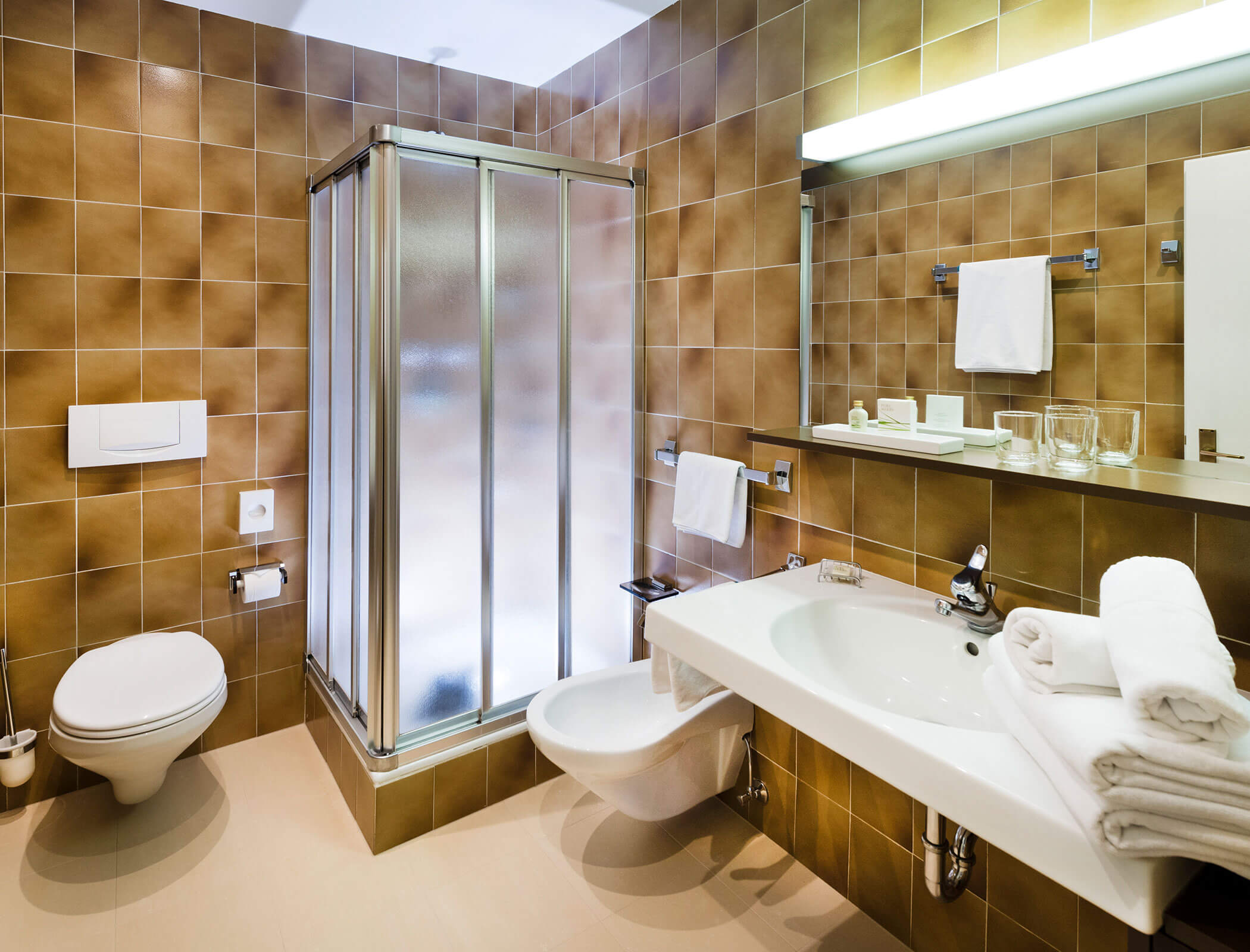 Modern bathroom with brown tiles, glass shower, toilet, sink, mirror, towels, and toiletries. - Hotel Fameli