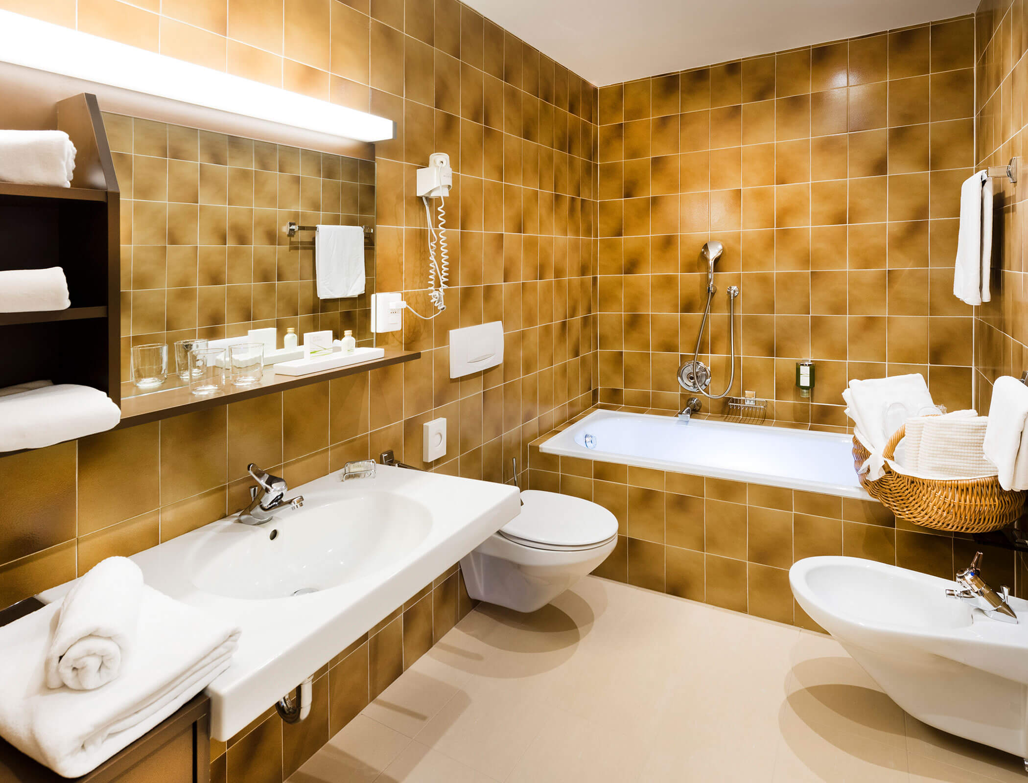 Modern bathroom with brown tiles, white sink, bathtub, toilet, bidet, towels, and shelves. - Hotel Fameli