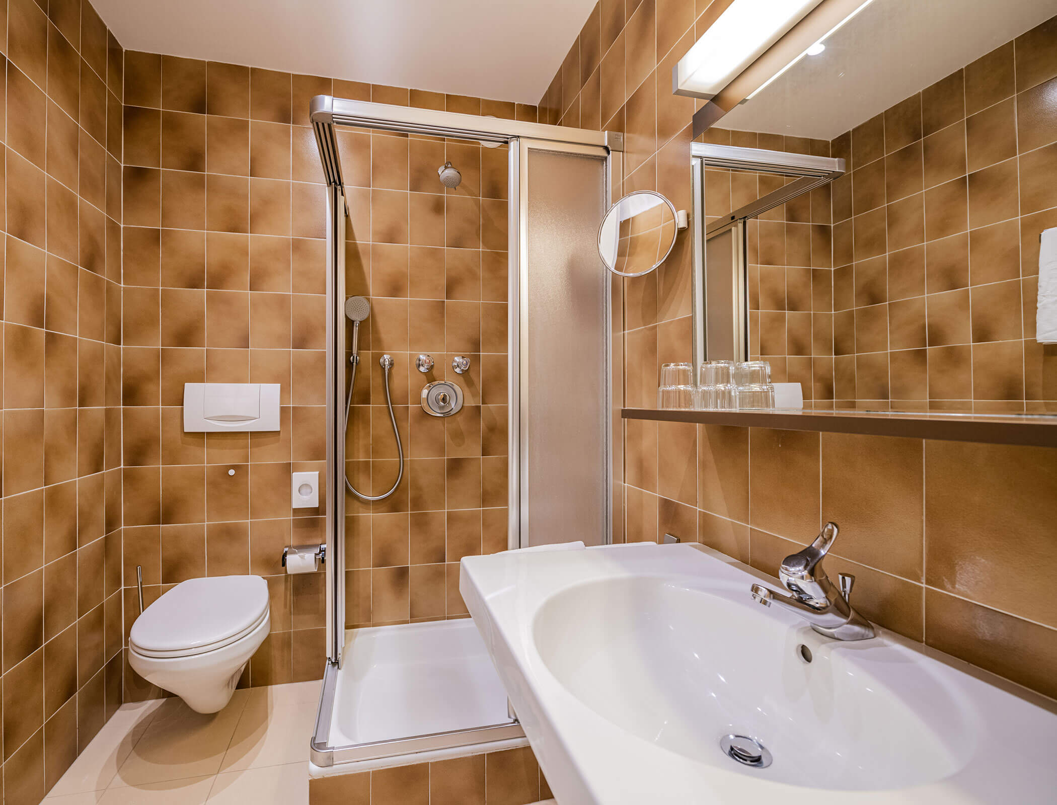 Bathroom with brown tiles, shower with glass door, white sink, mirror, and wall-mounted toilet. - Hotel Fameli
