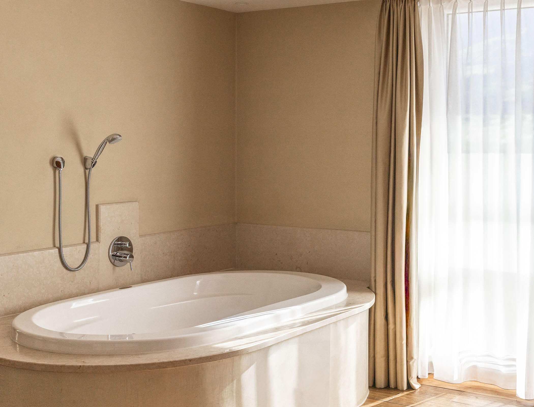 Sunlit bathroom with a white oval bathtub, handheld shower, beige walls, and sheer curtains. - Hotel Fameli