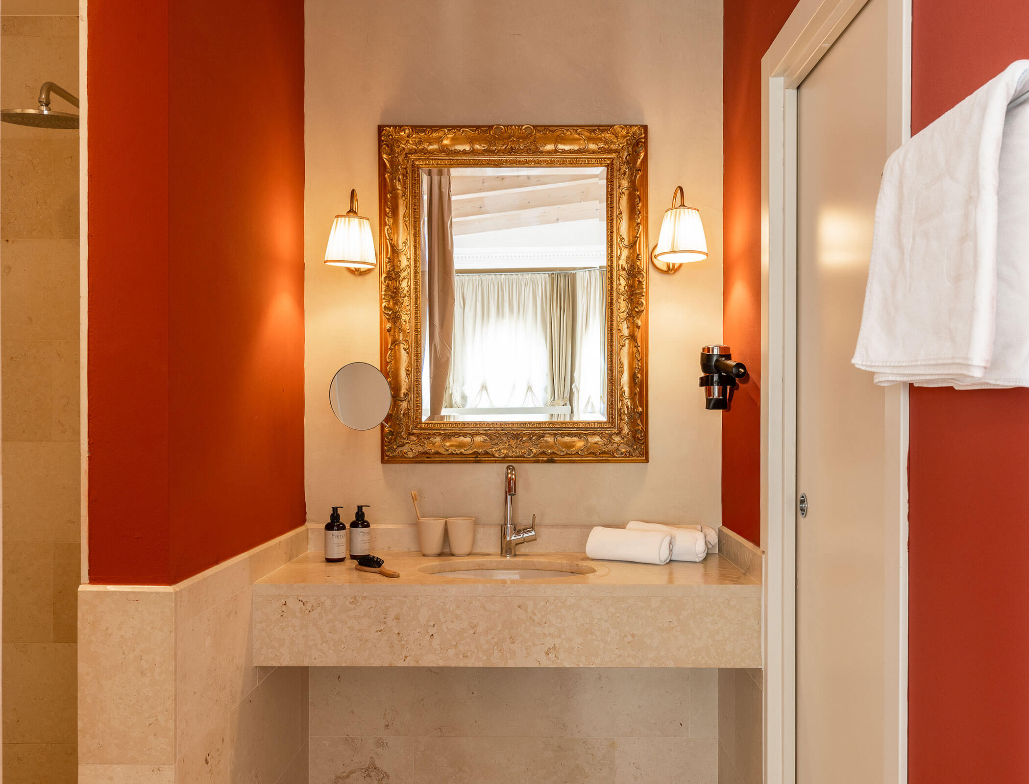 Elegant bathroom vanity with a gold-framed mirror, lights, towels, and toiletries on a beige countertop. - Hotel Fameli