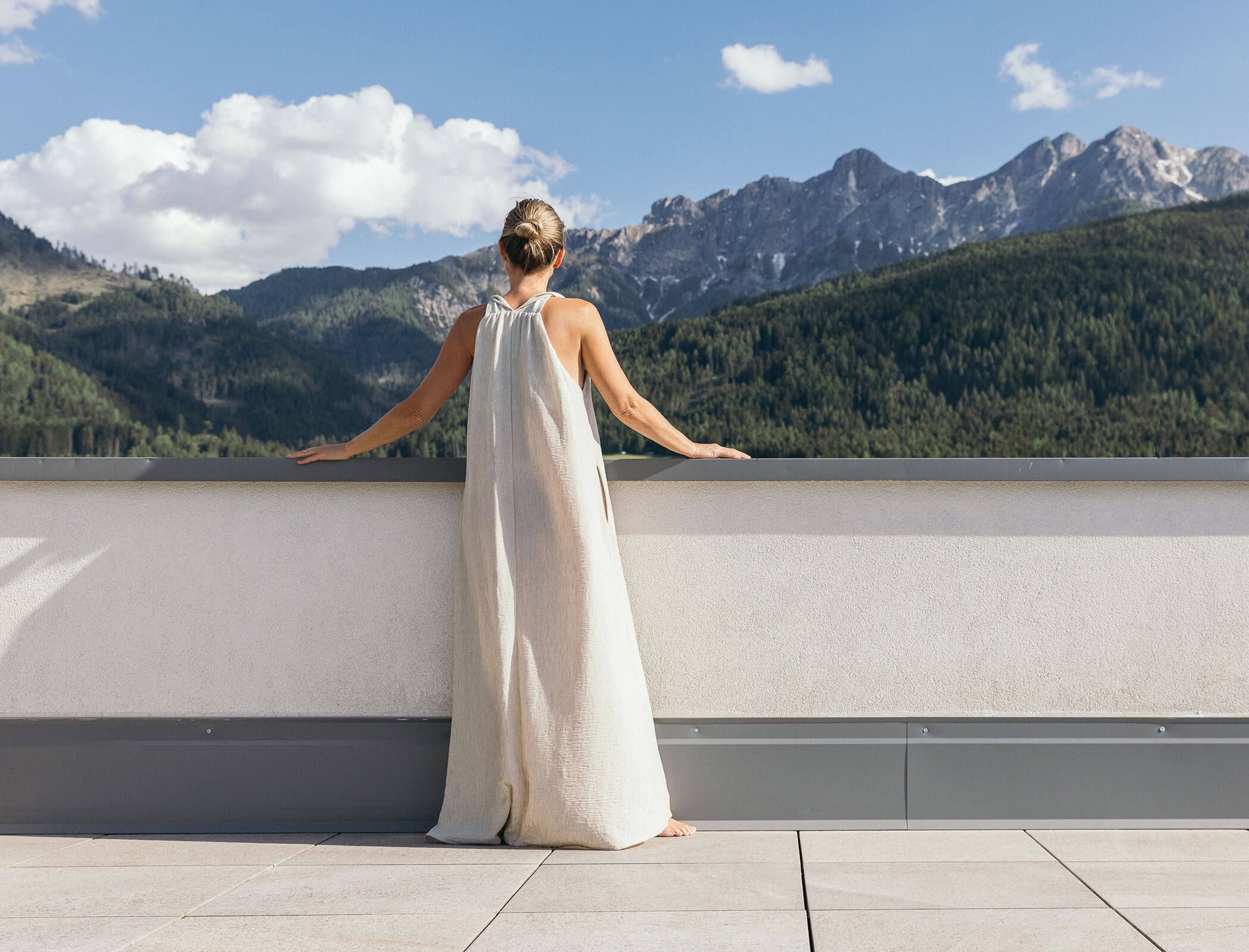 Woman in a long dress stands on a terrace, looking at mountains under a blue sky with clouds. - Hotel Fameli