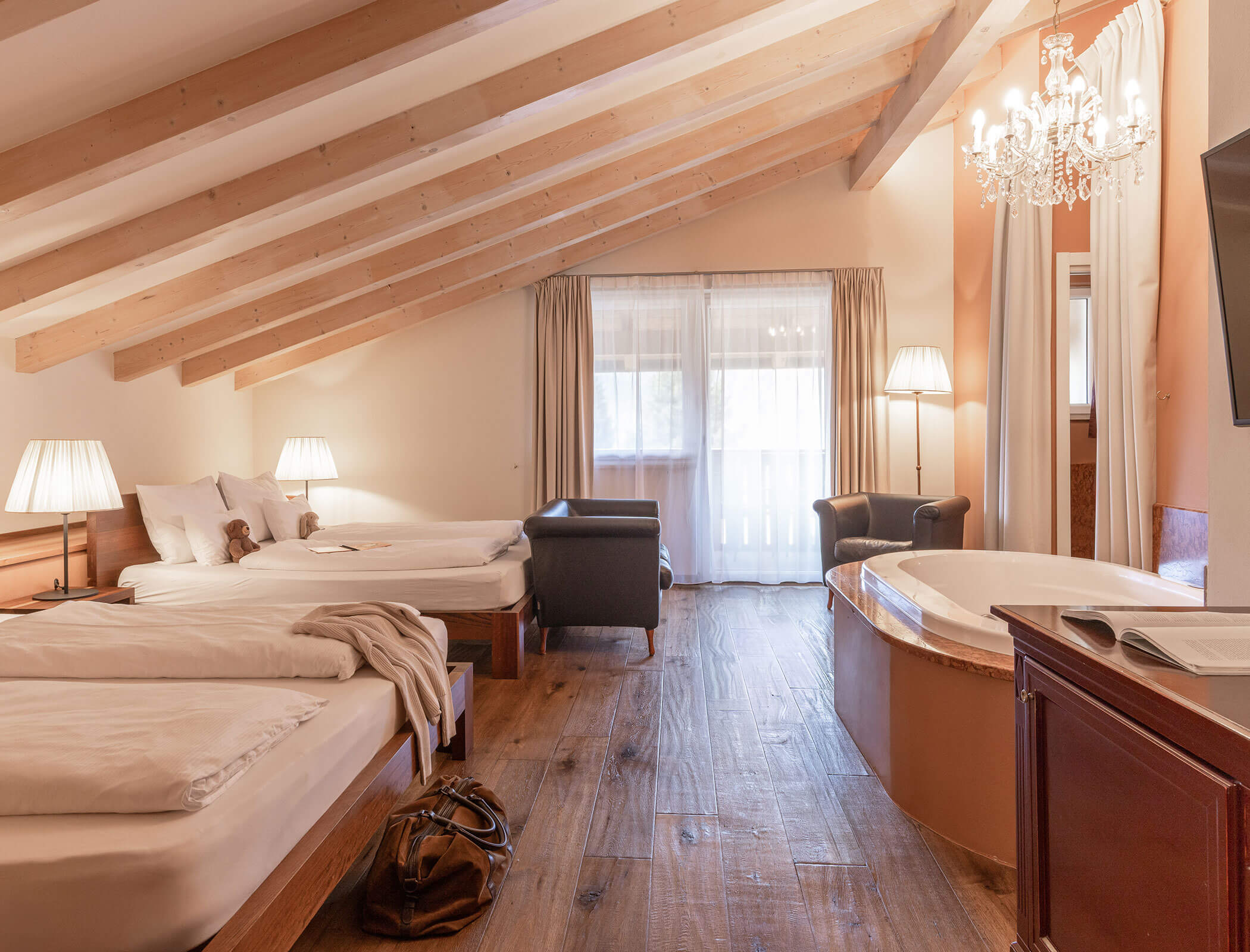 Spacious attic hotel room with two beds, a hot tub, armchairs, and wooden floors under a chandelier. - Hotel Fameli