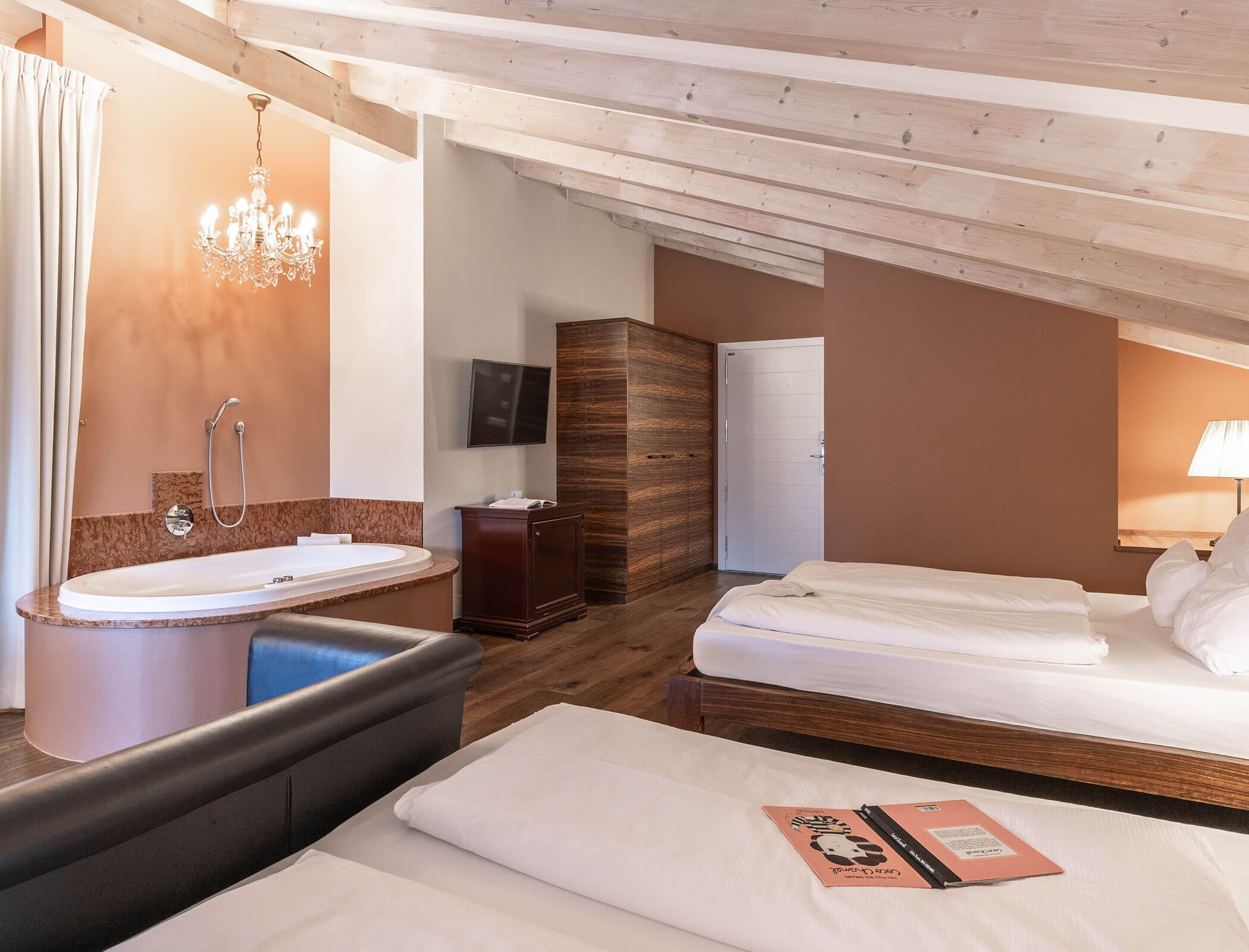 Cozy attic hotel room with two beds, a chandelier, a bathtub, TV, and wood-paneled furniture. - Hotel Fameli