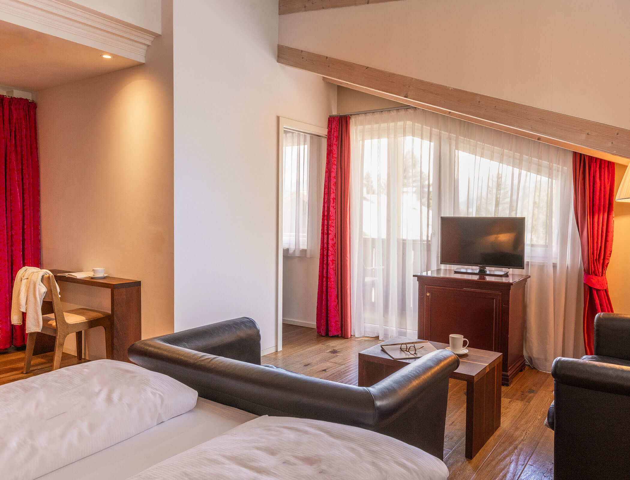 Cozy hotel room with a bed, desk, chairs, TV, red curtains, and wooden floors, softly lit by sunlight. - Hotel Fameli