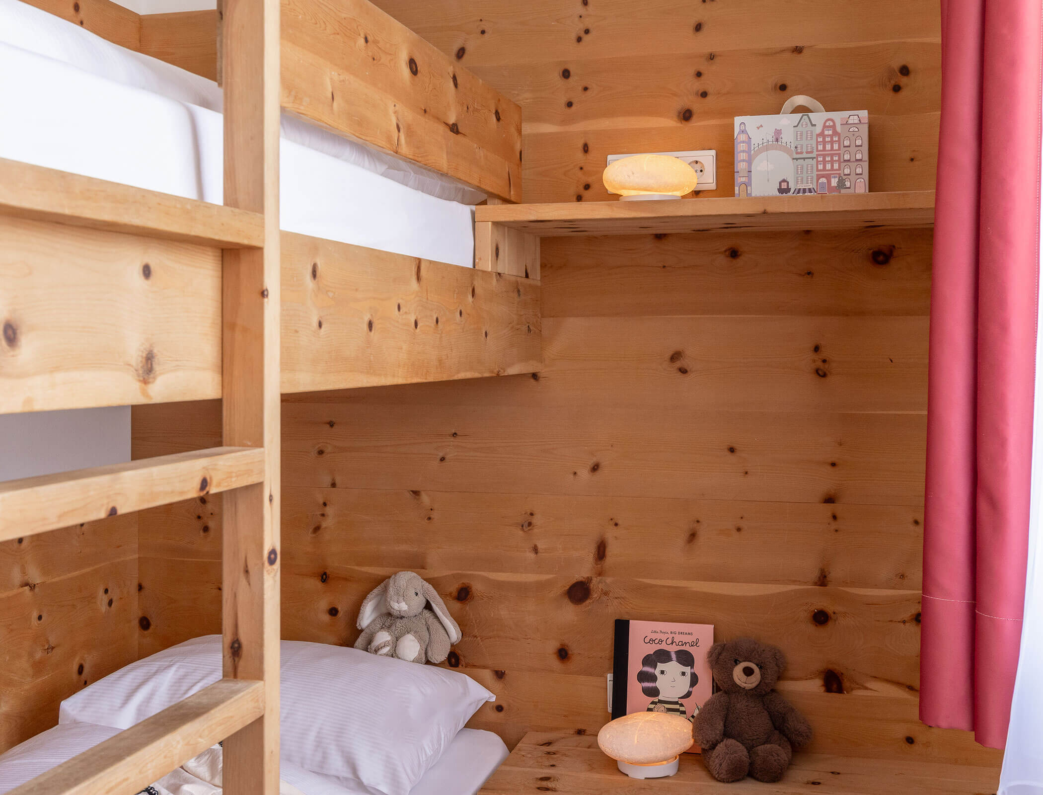 Wooden bunk bed with stuffed animals, books, and a lamp on a shelf, next to a pink curtain. - Hotel Fameli