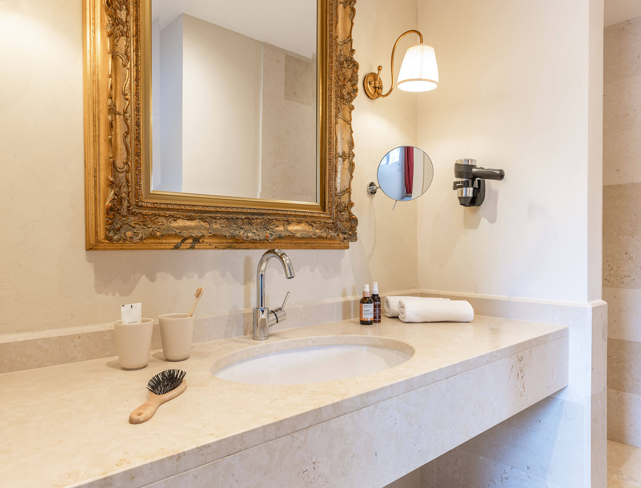 Elegant bathroom vanity with a gold-framed mirror, toiletries, towel, and brush on a beige countertop. - Hotel Fameli