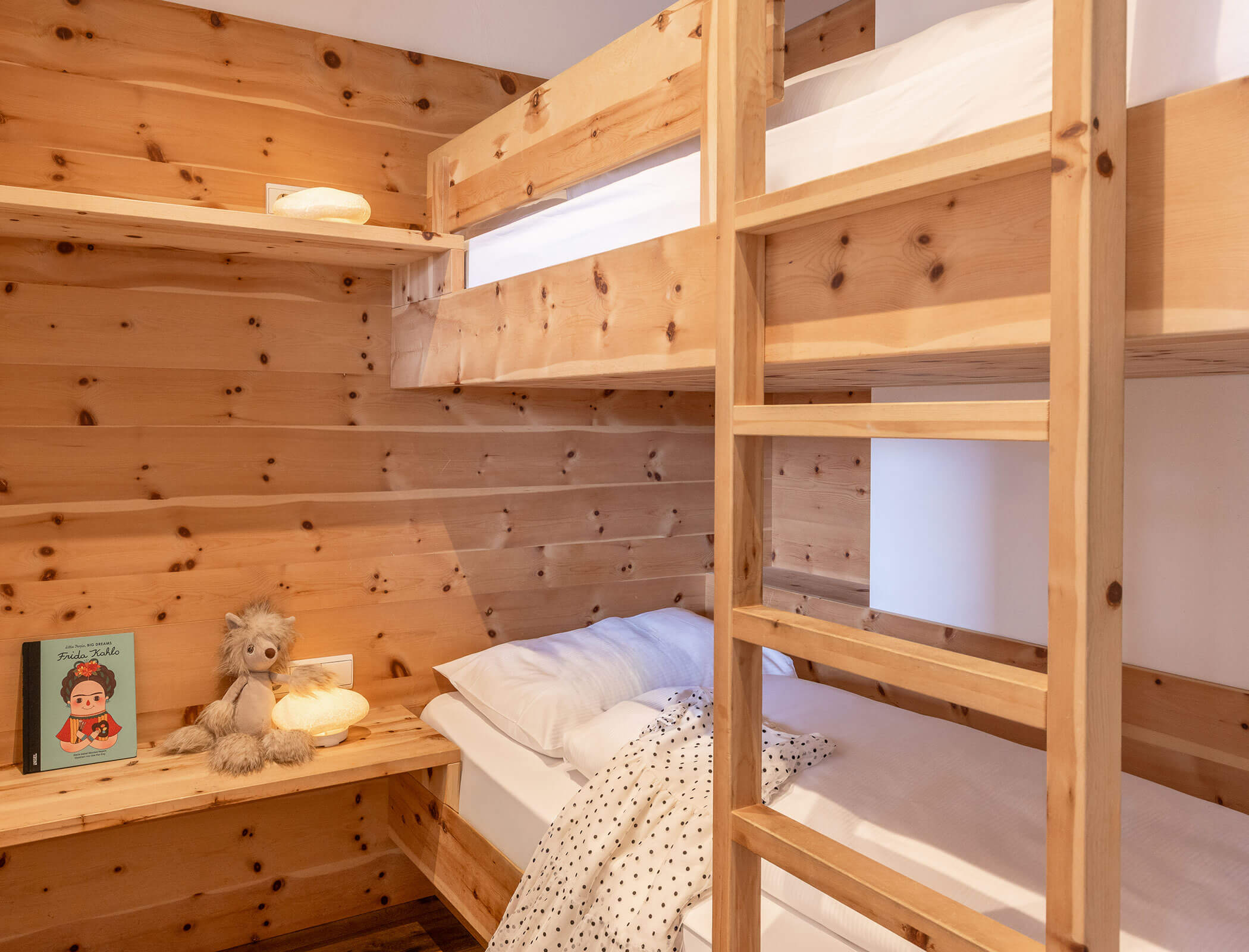 Wooden bunk beds in a cozy room with shelves, a teddy bear, a book, and a dotted blanket. - Hotel Fameli