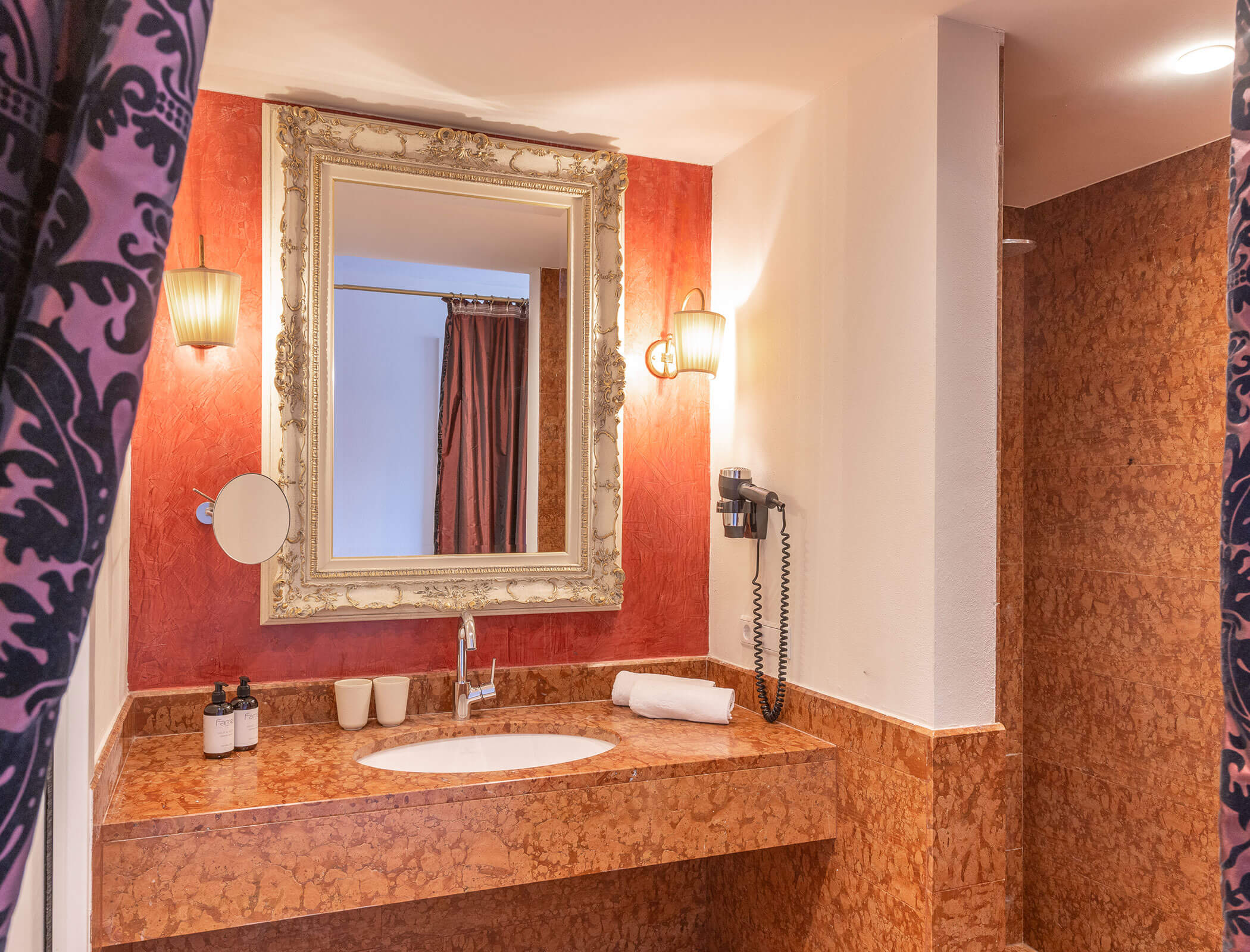 Bathroom with ornate mirror, marble counter, and wall sconces, with a towel and toiletries on the sink. - Hotel Fameli