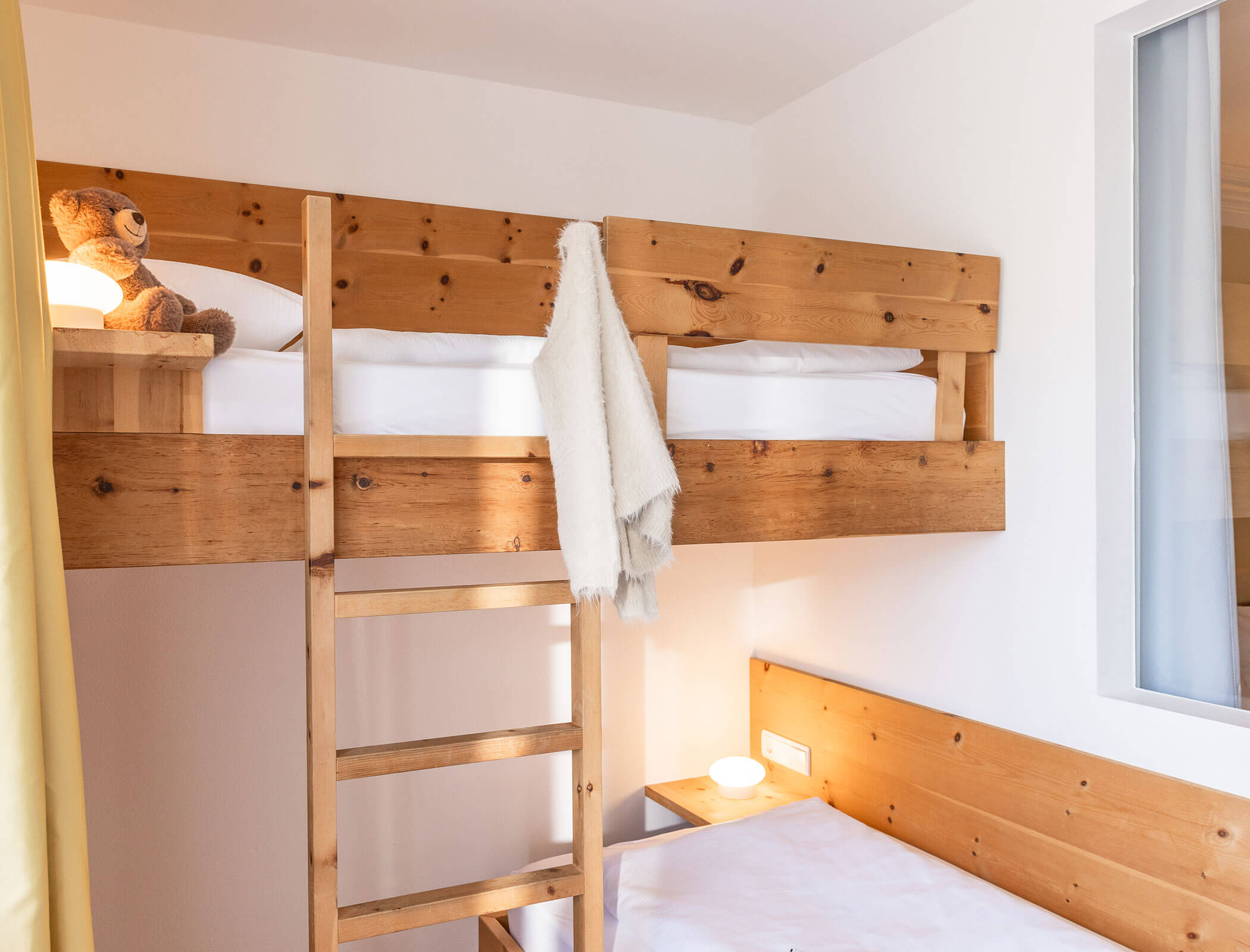 Wooden bunk bed with a teddy bear, white blanket, and ladder, next to a white wall and window. - Hotel Fameli