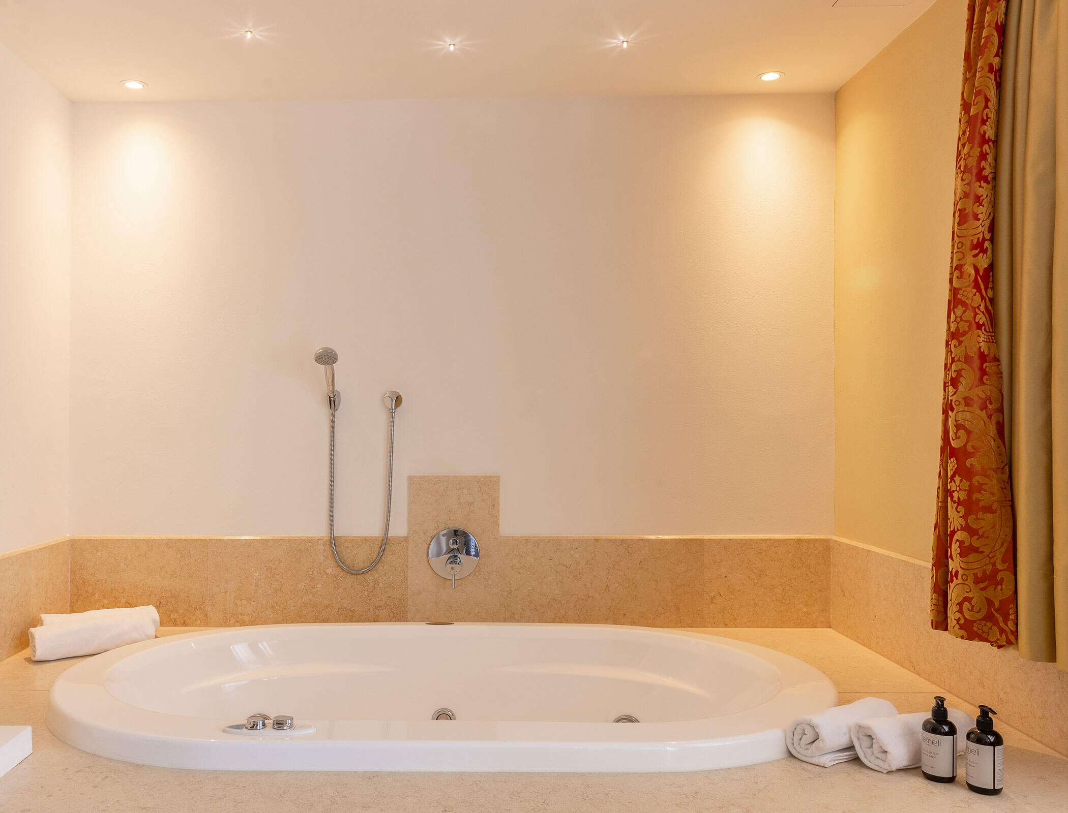 Large white bathtub with towels, toiletries, wall-mounted shower, and warm lighting in a beige bathroom. - Hotel Fameli