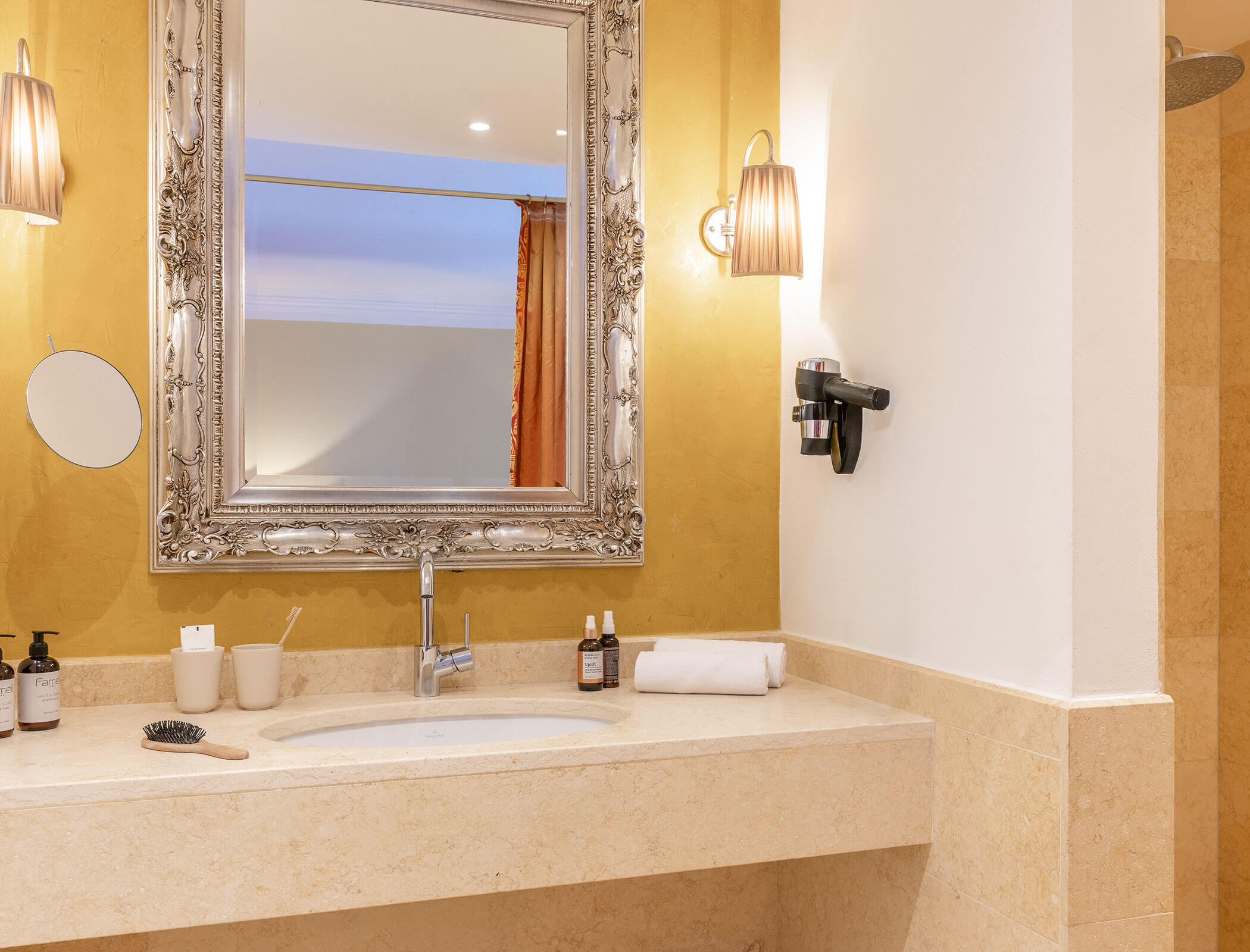 Elegant bathroom with ornate mirror, wall lights, sink, toiletries, towel, and beige counter. - Hotel Fameli