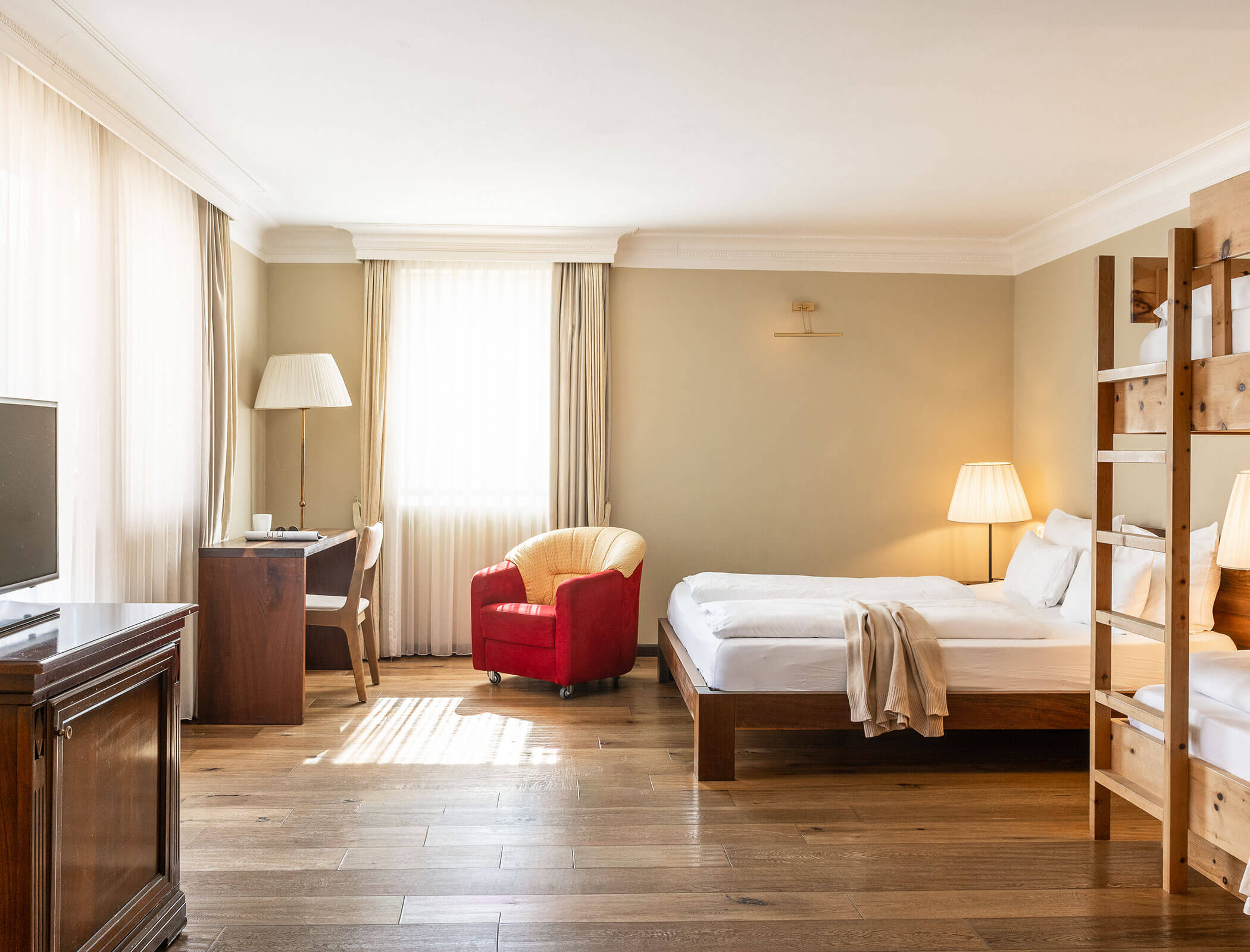Bright hotel room with a bed, desk, red chair, TV, and wooden floors, lit by natural light. - Hotel Fameli