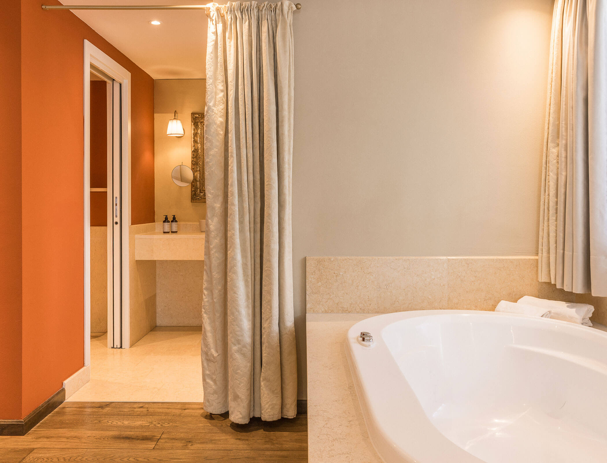 Modern bathroom with a white bathtub, tan walls, curtain, and a view into a vanity area with a mirror. - Hotel Fameli