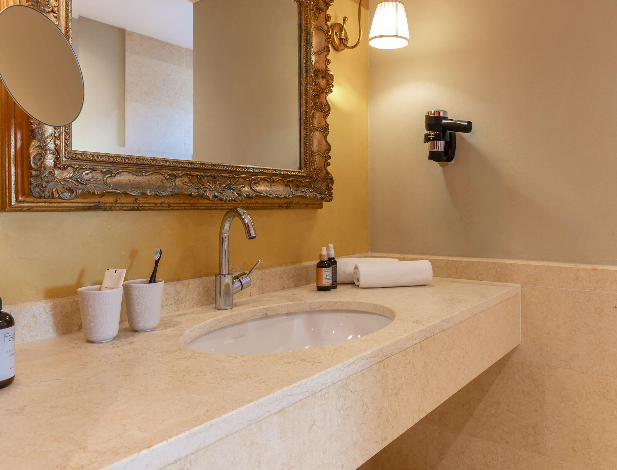 Elegant bathroom sink with ornate mirror, towels, toiletries, and wall-mounted hair dryer. - Hotel Fameli