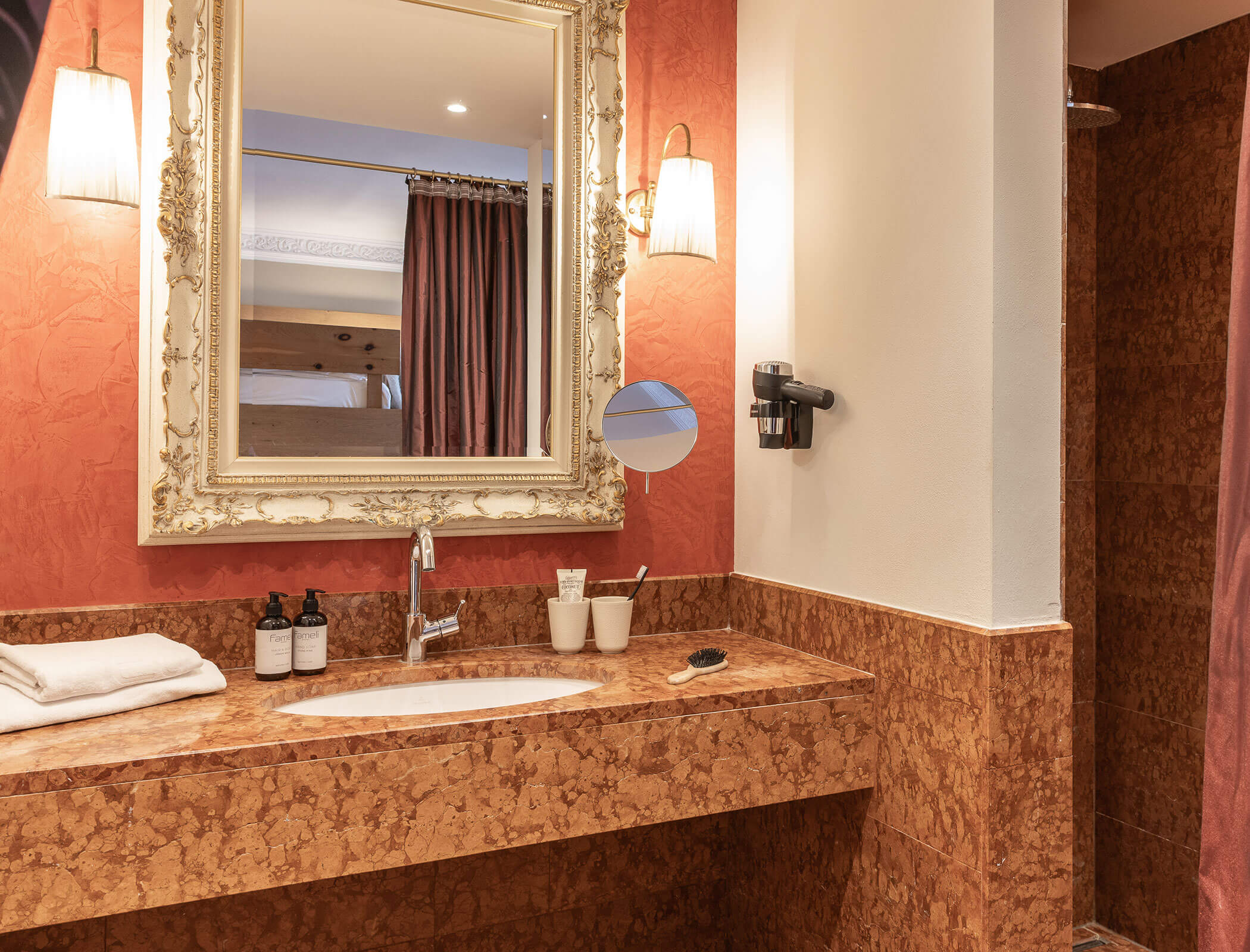 Bathroom with brown marble sink, large mirror, towels, toiletries, wall lights, and a shower area. - Hotel Fameli