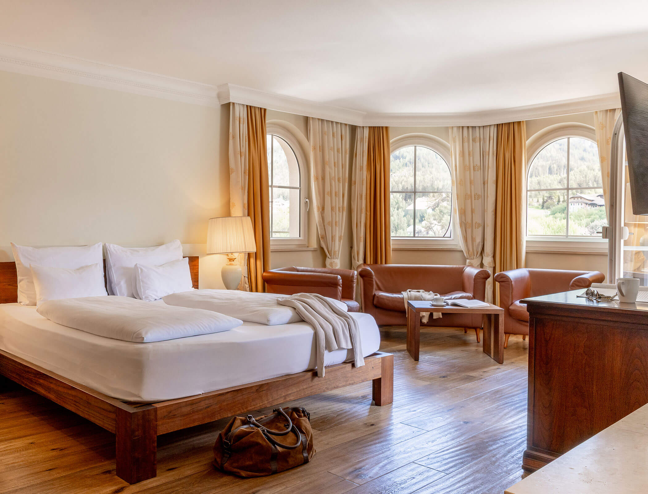 Bright hotel room with a wooden bed, leather chairs, large windows, and a view of trees outside. - Hotel Fameli