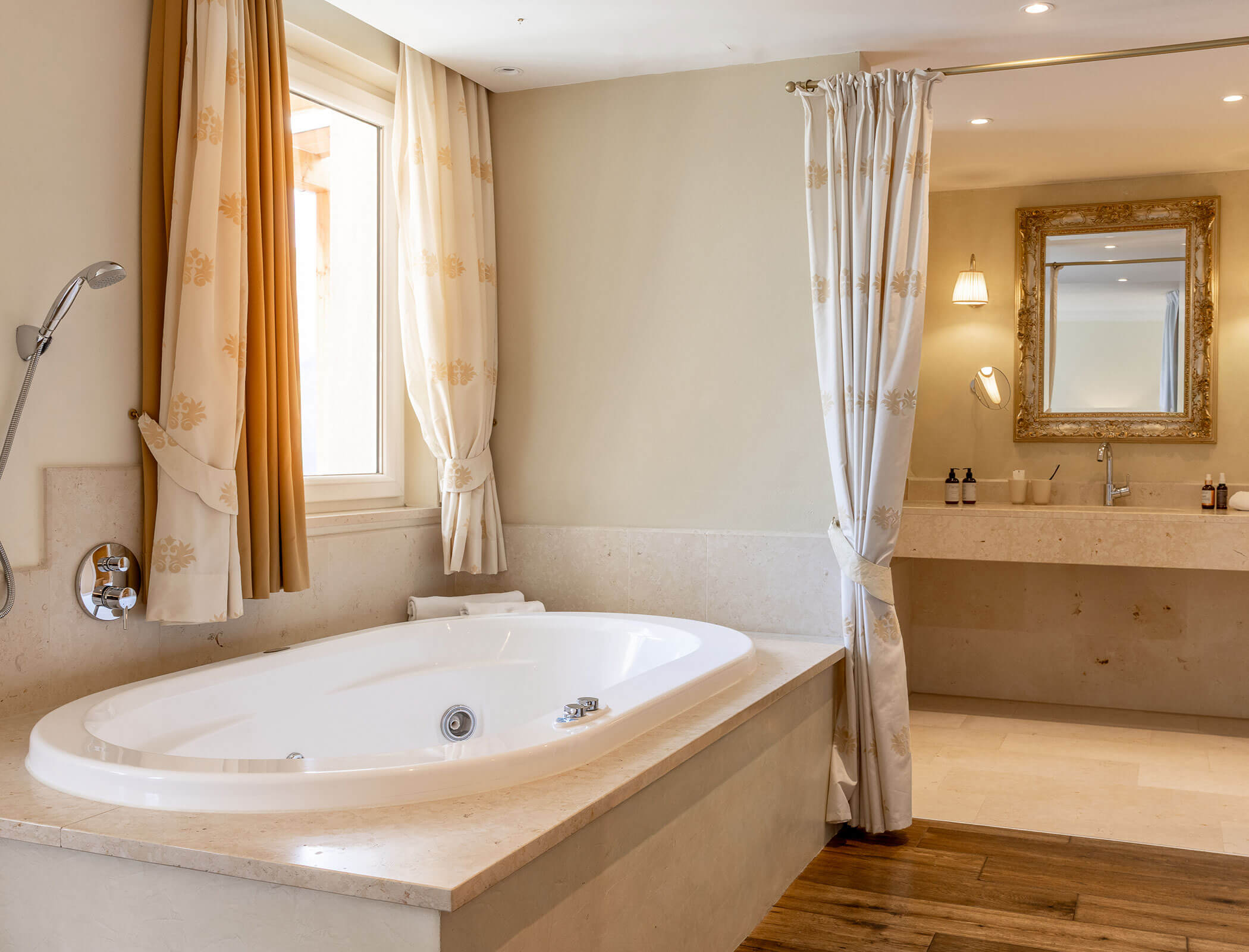 Spacious bathroom with a bathtub, window, curtains, large mirror, sink, and wooden floor accents. - Hotel Fameli