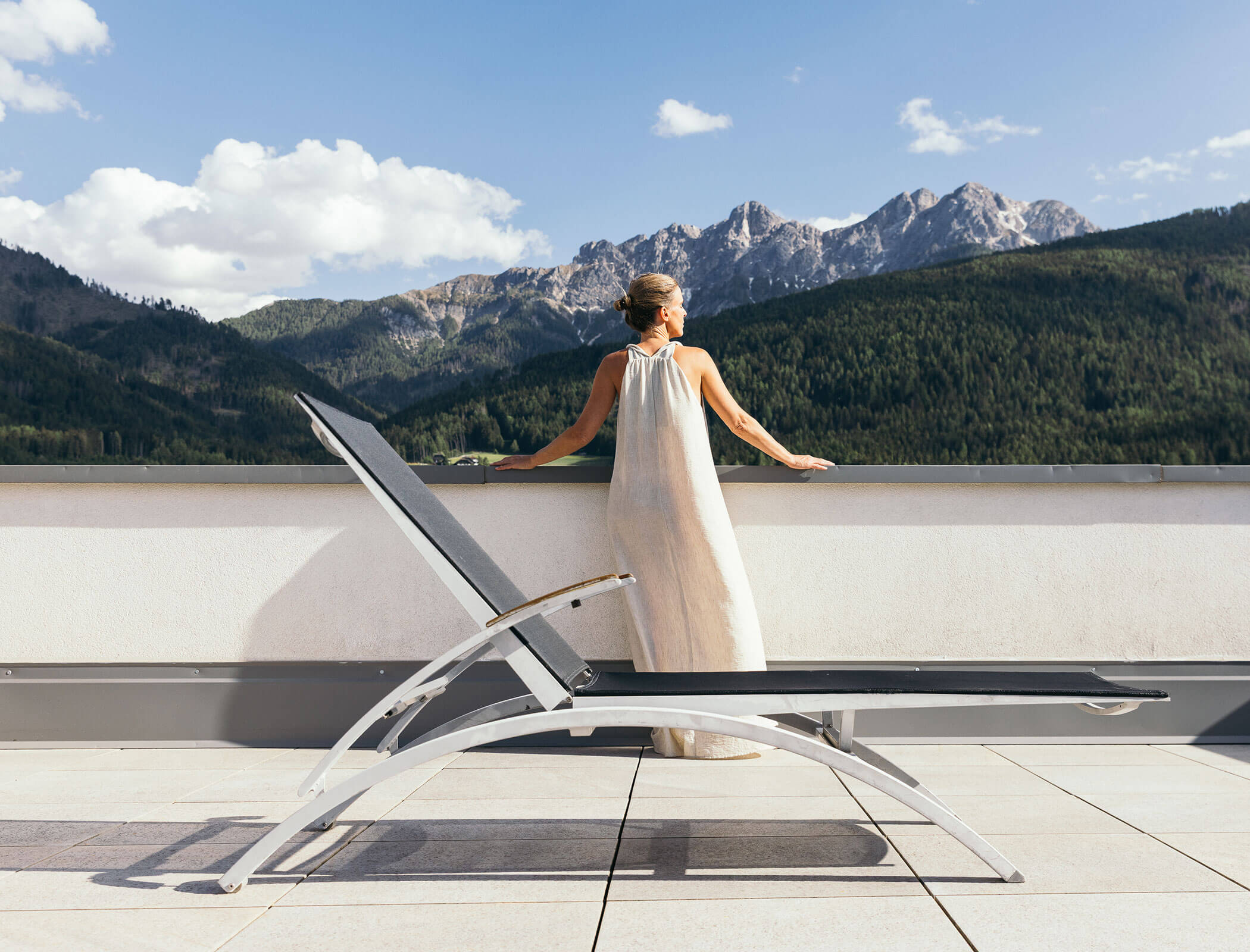 Woman in a long dress stands by a railing, overlooking mountains and forest beside an empty lounge chair. - Hotel Fameli