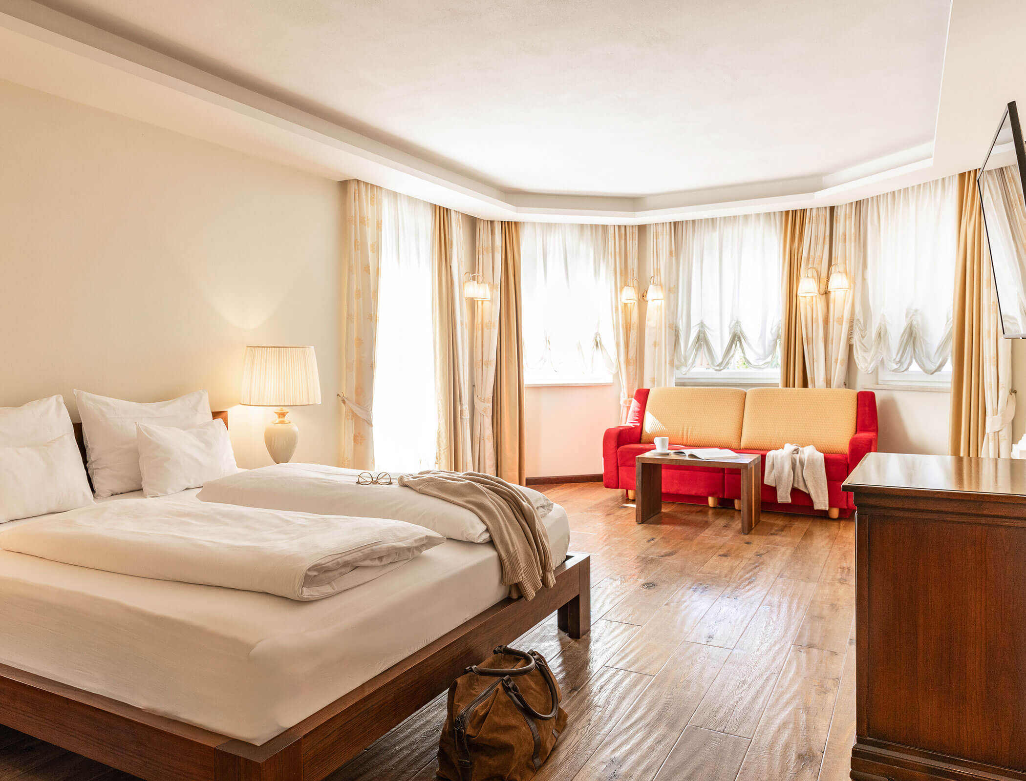 Bright hotel room with a large bed, red sofa, coffee table, and sunlight streaming through windows. - Hotel Fameli