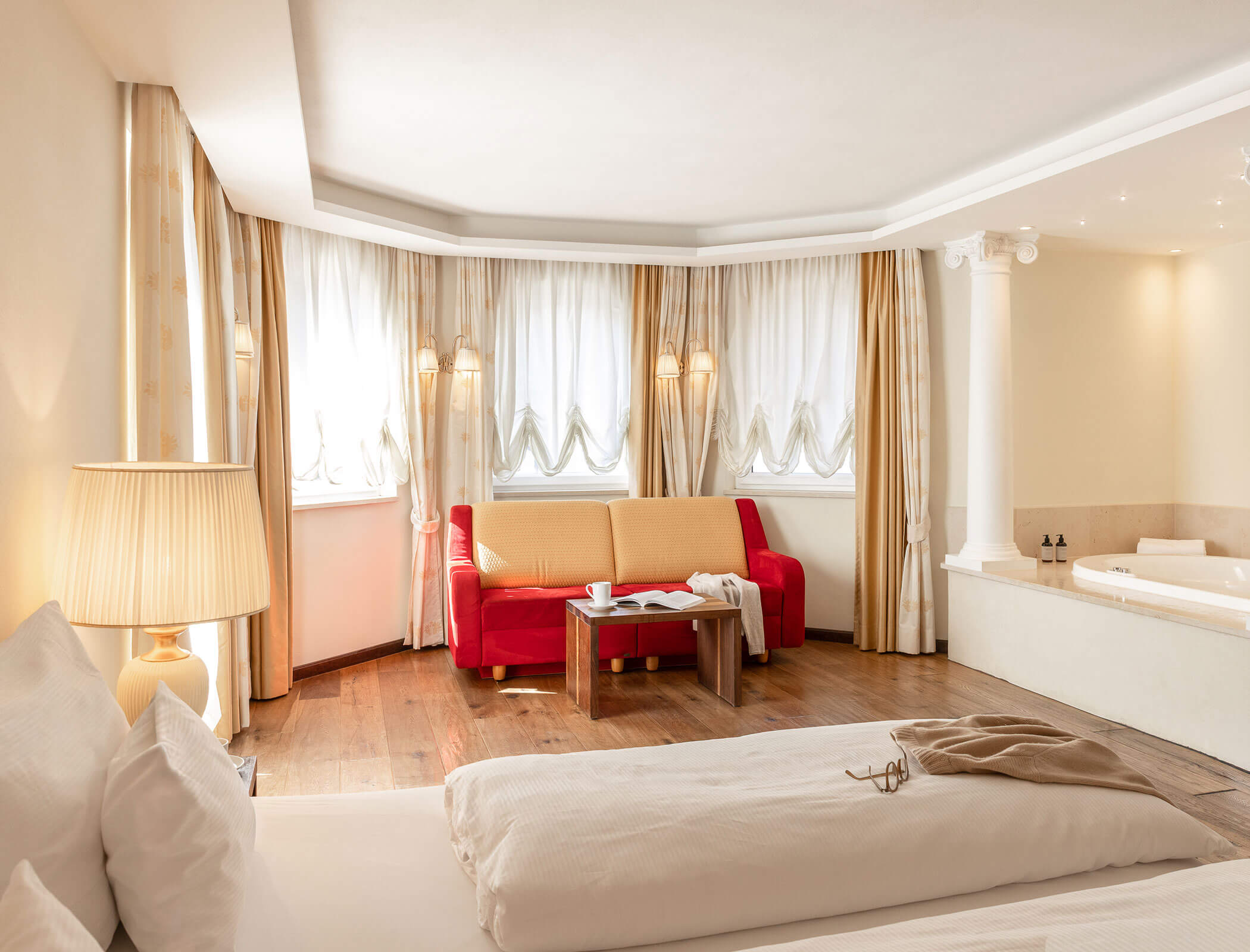 Bright hotel room with bed, red sofa, table, large windows, and corner bathtub, decorated in neutral tones. - Hotel Fameli
