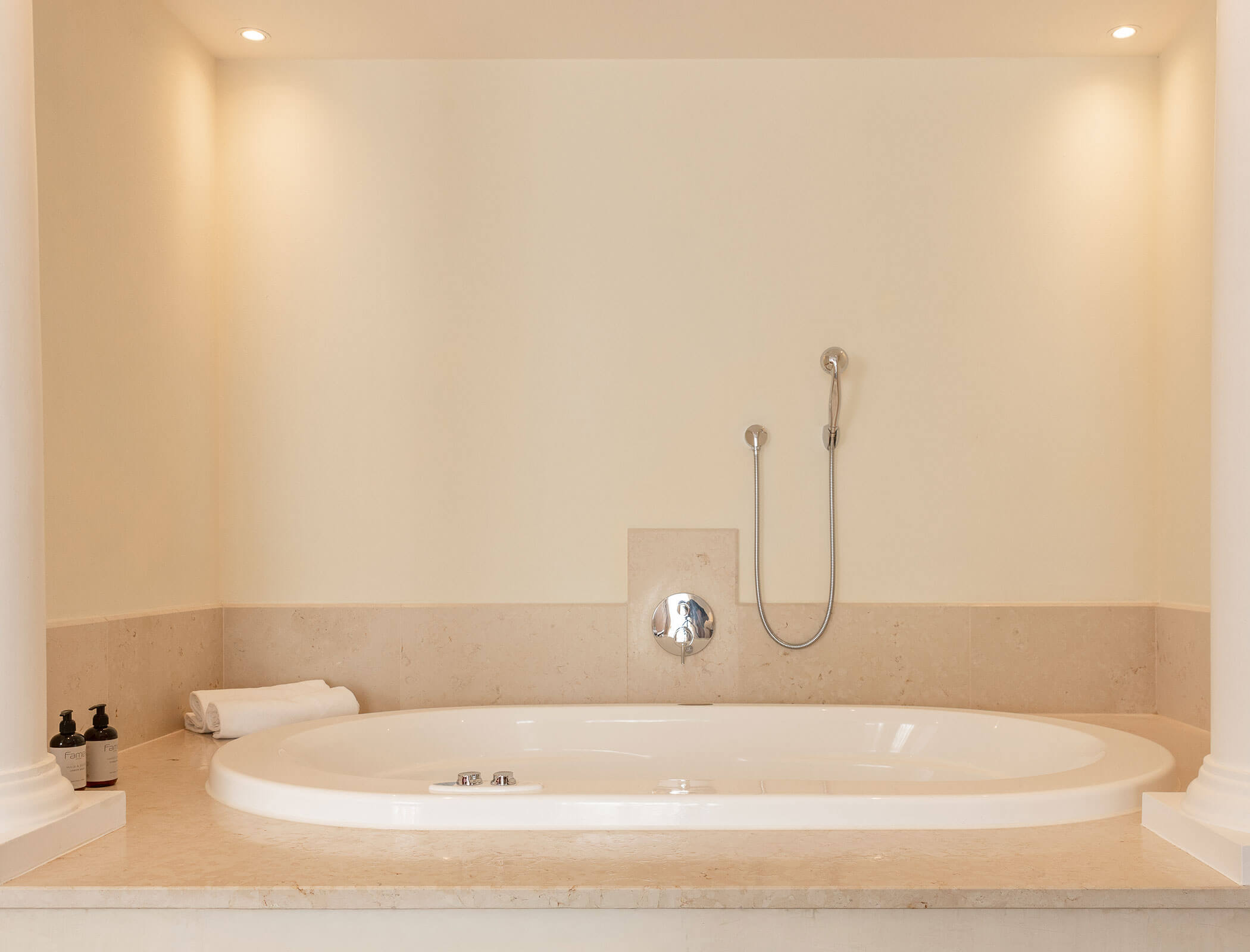 A modern built-in bathtub with a towel, bath products, and a handheld shower against a beige wall. - Hotel Fameli