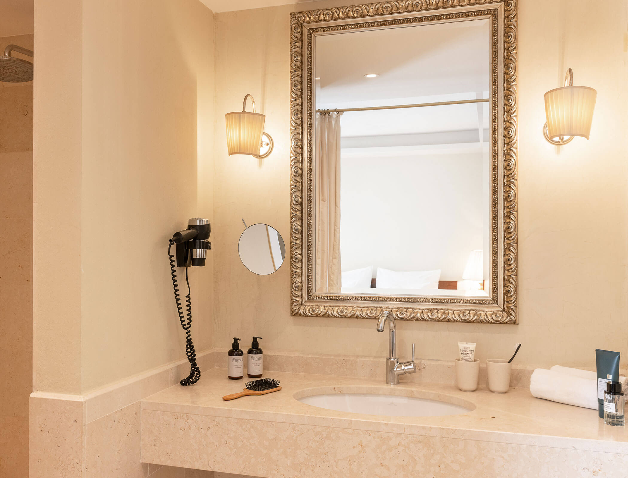 Bathroom vanity with a large mirror, wall lights, sink, toiletries, and a mounted hairdryer. - Hotel Fameli