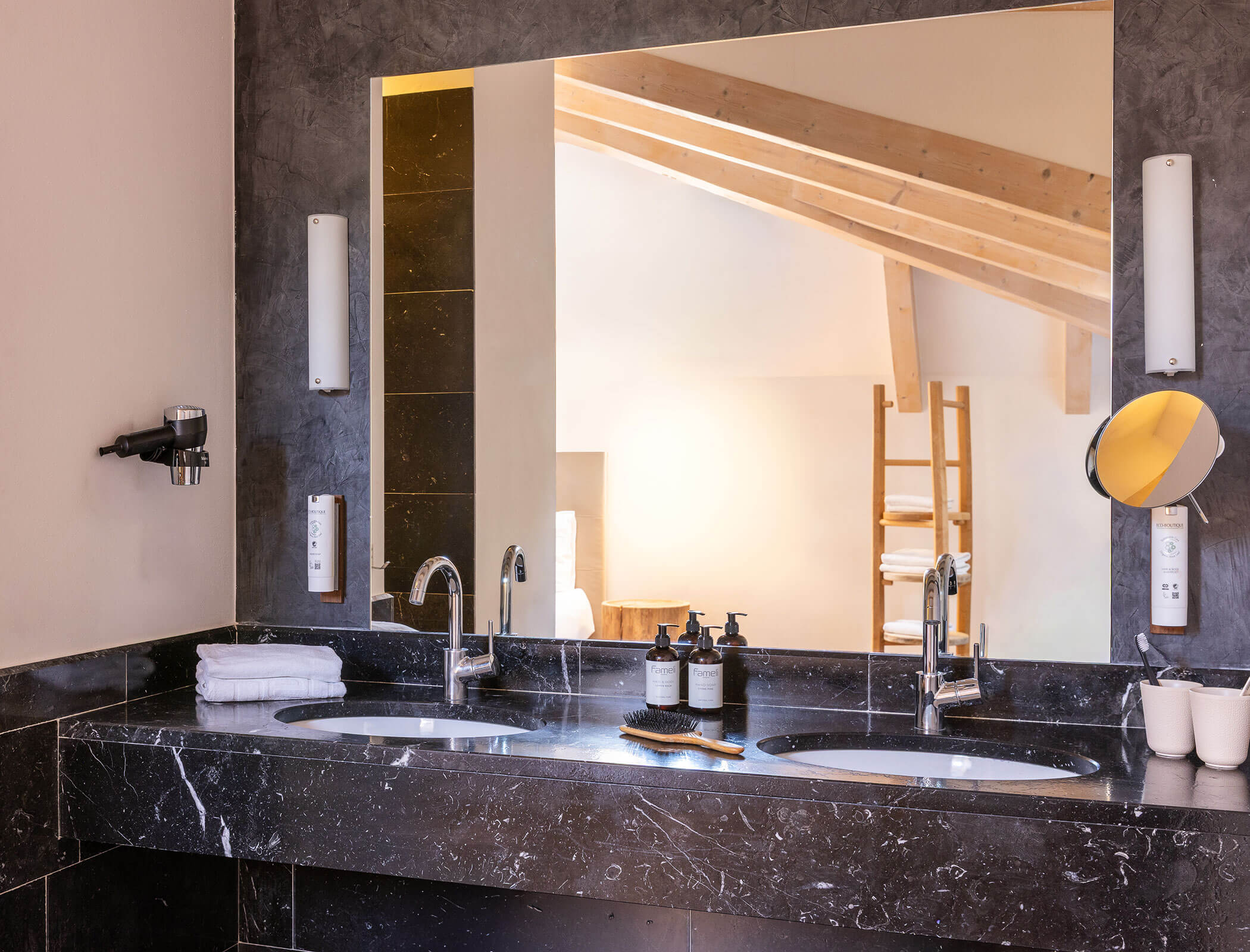 Double sink bathroom with marble countertop, mirror, toiletries, towels, and wooden beams reflected. - Hotel Fameli