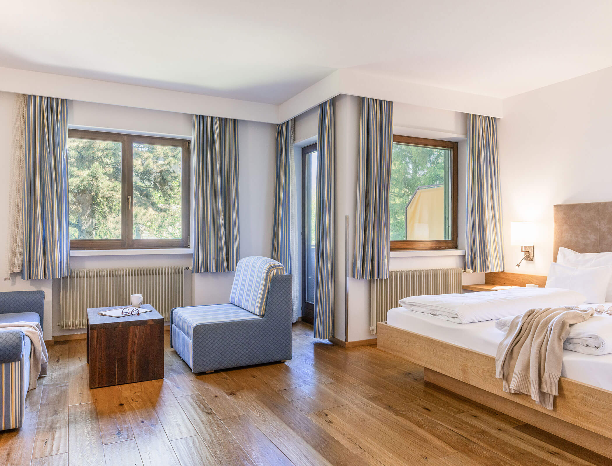 Bright hotel room with large windows, double bed, blue chairs, wooden floor, and light curtains. - Hotel Fameli