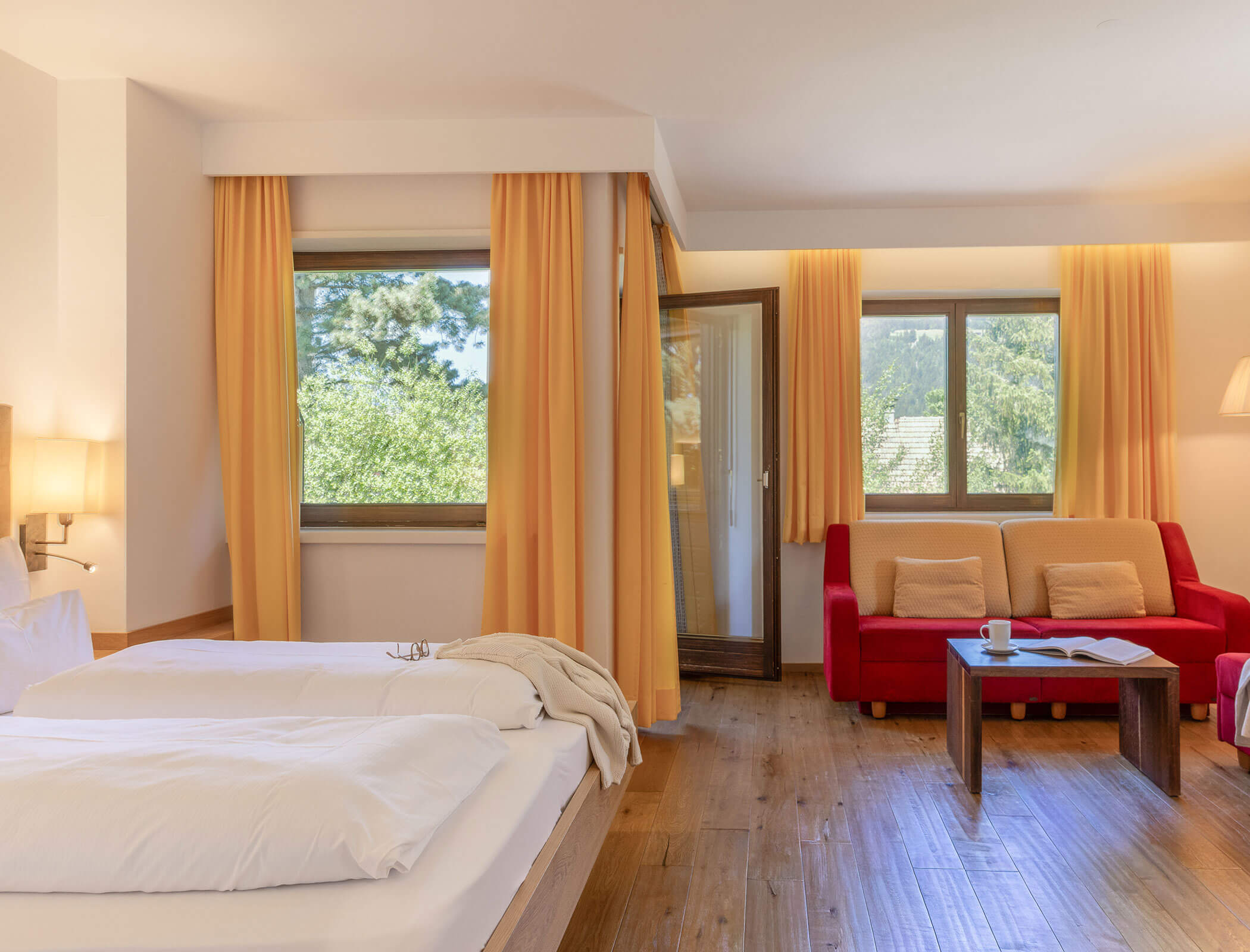 Bright hotel room with a double bed, red sofa, large windows, wooden floor, and yellow curtains. - Hotel Fameli