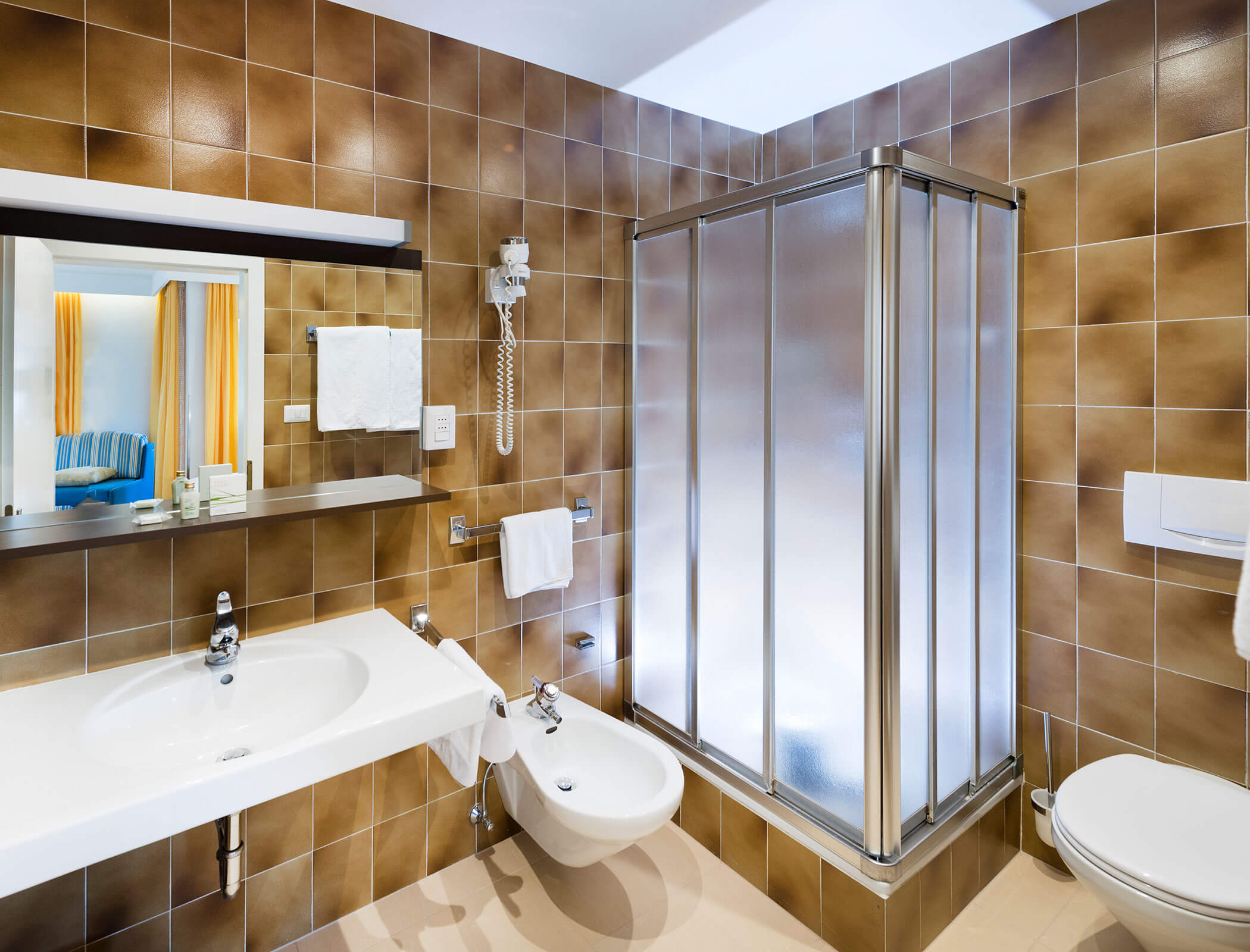 Modern bathroom with brown tiles, glass shower, sink, bidet, toilet, mirror, and towel racks. - Hotel Fameli