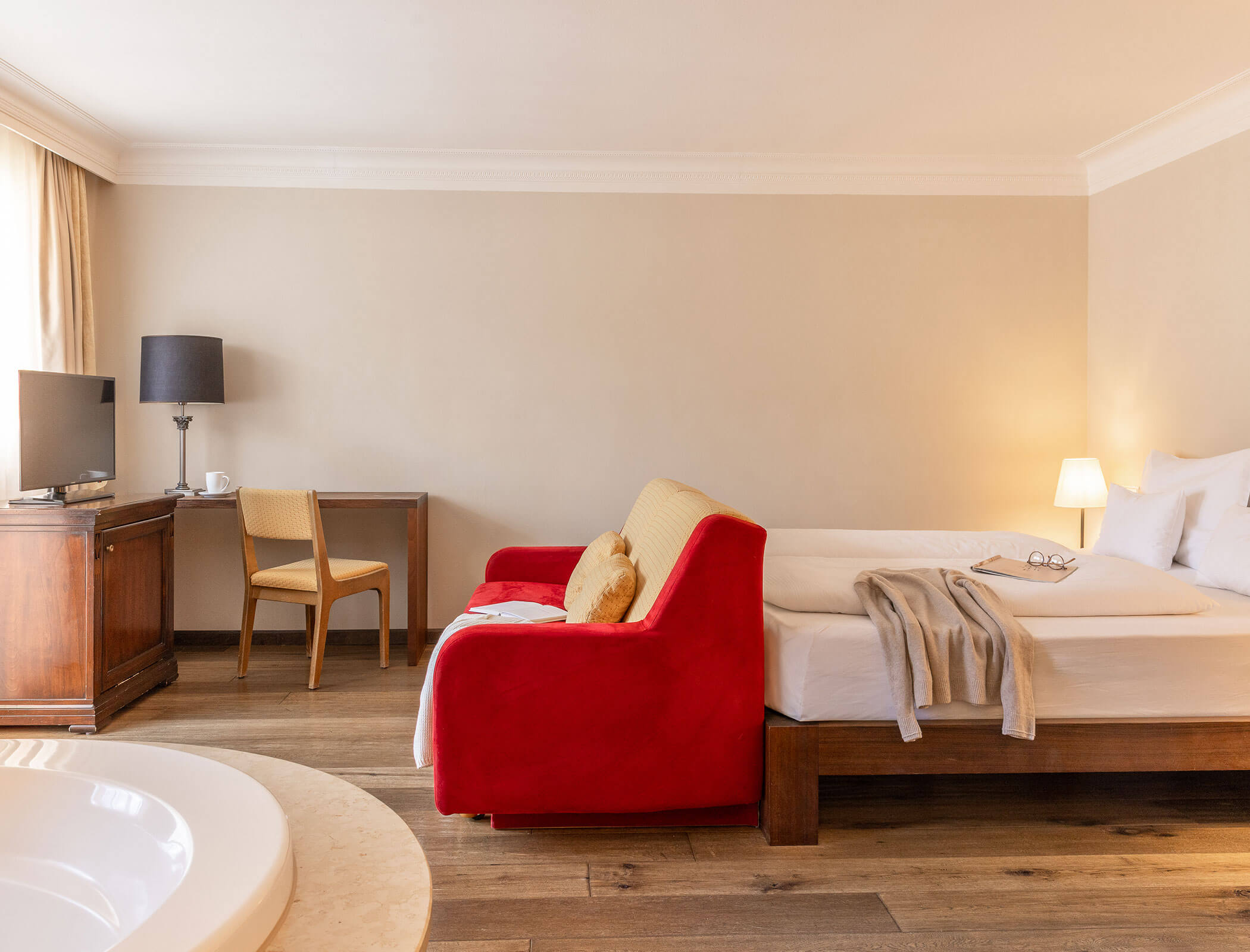 Hotel room with a red sofa, bed, desk, TV, lamps, and a round tub in the foreground. - Hotel Fameli