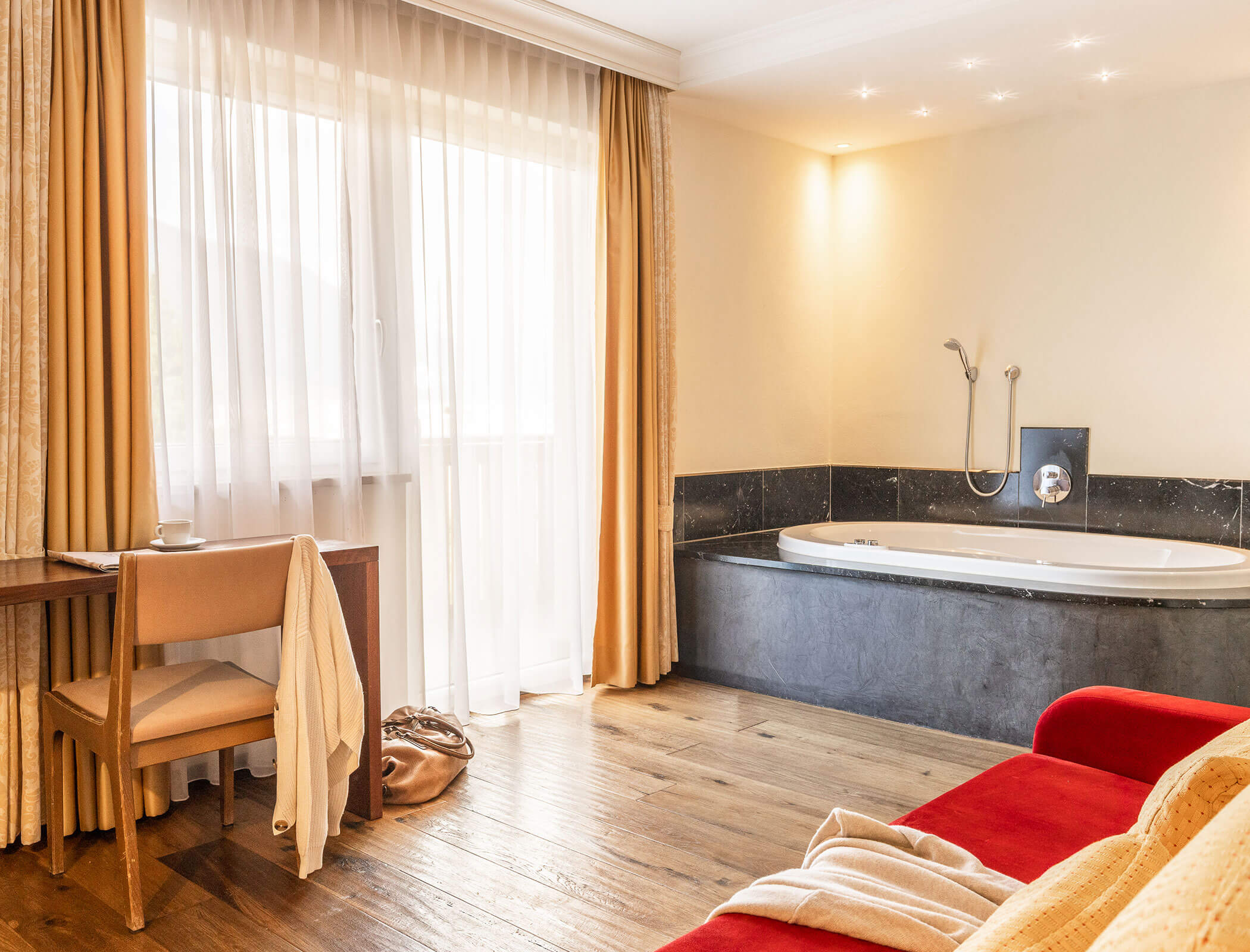 A hotel room with a desk, chair, sofa, window with curtains, and a bathtub in the corner. - Hotel Fameli