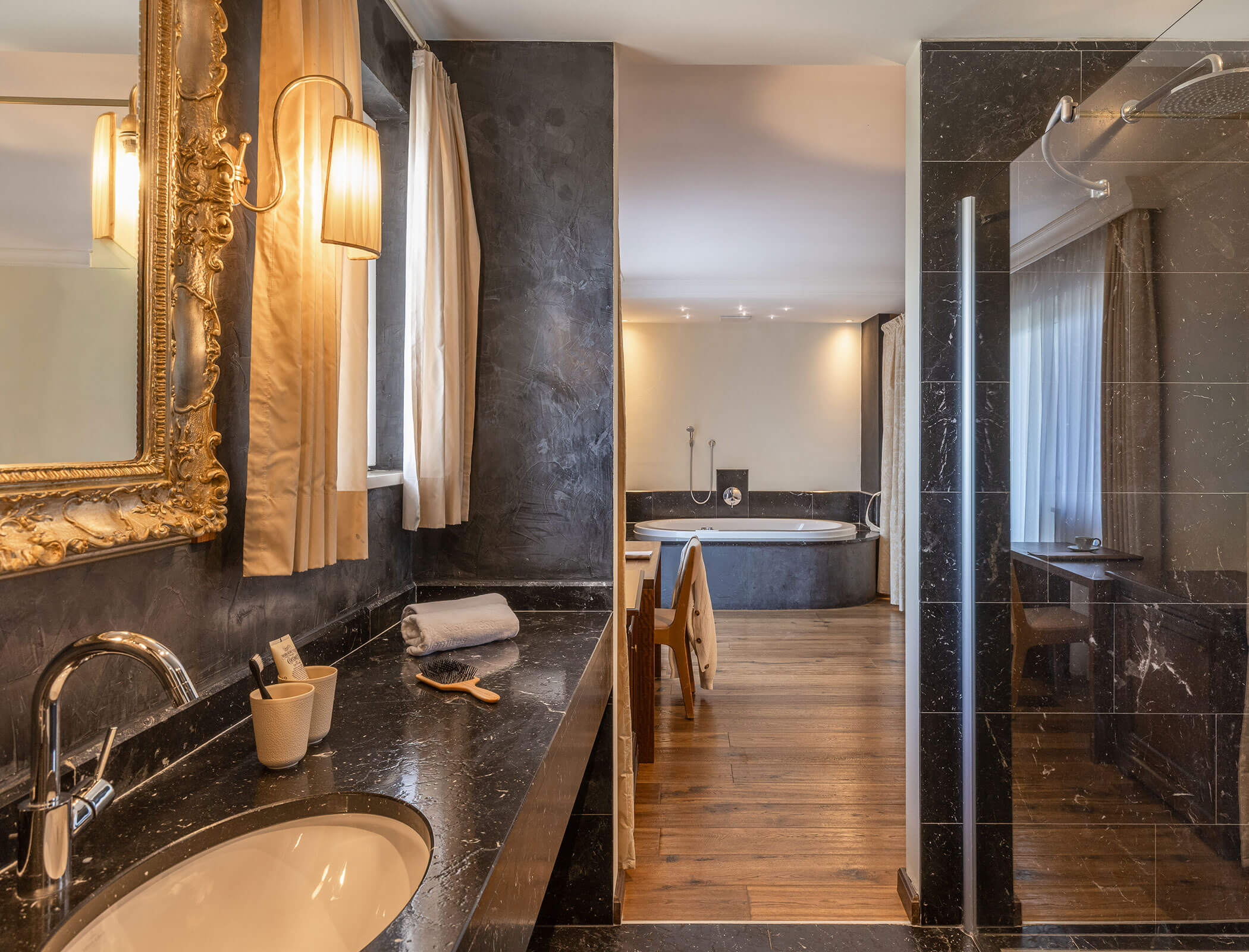 Elegant bathroom with a black marble sink, large mirror, walk-in shower, and soaking tub in the background. - Hotel Fameli