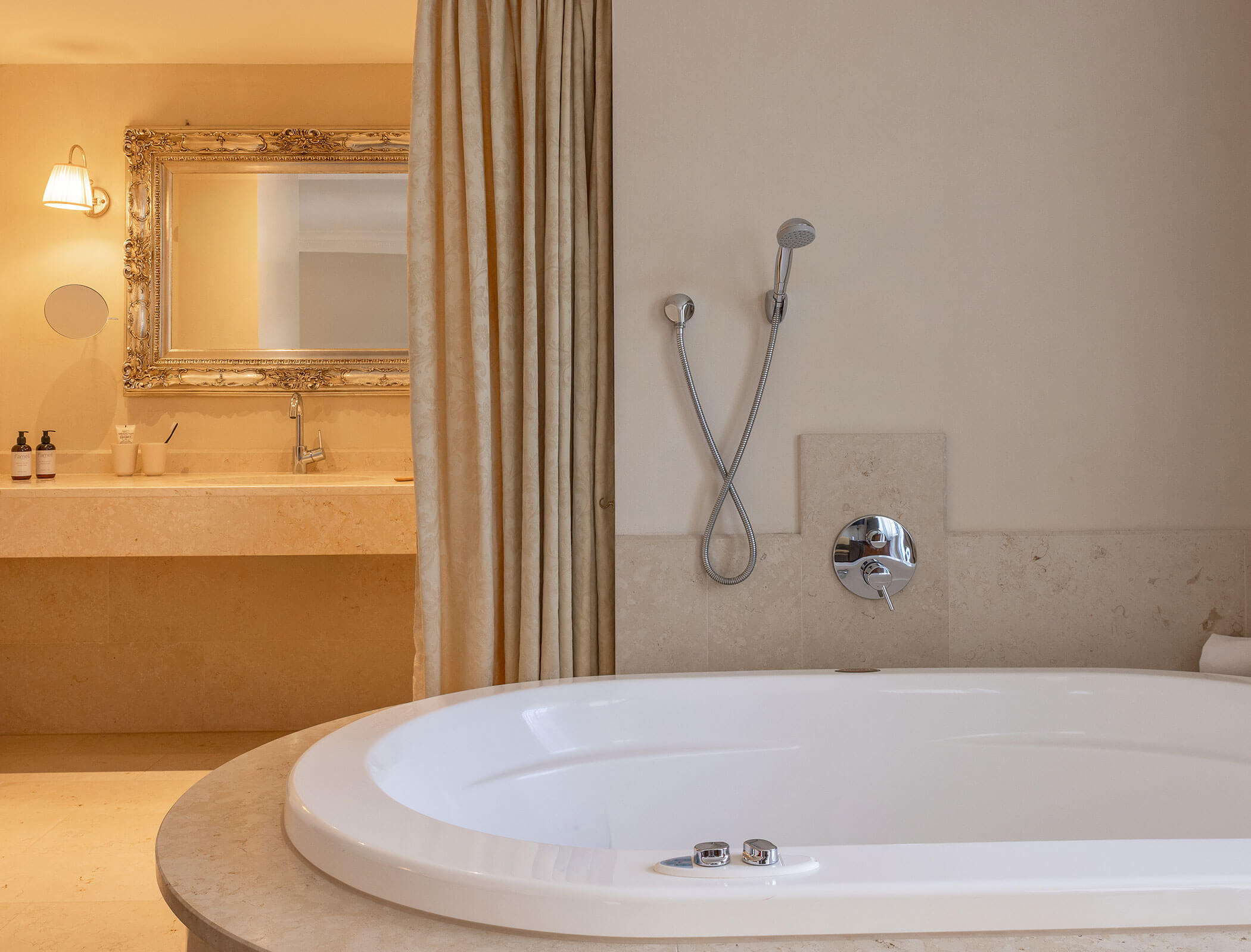 Oval bathtub with handheld shower, beige walls, curtain, and ornate mirror above a sink in bathroom. - Hotel Fameli