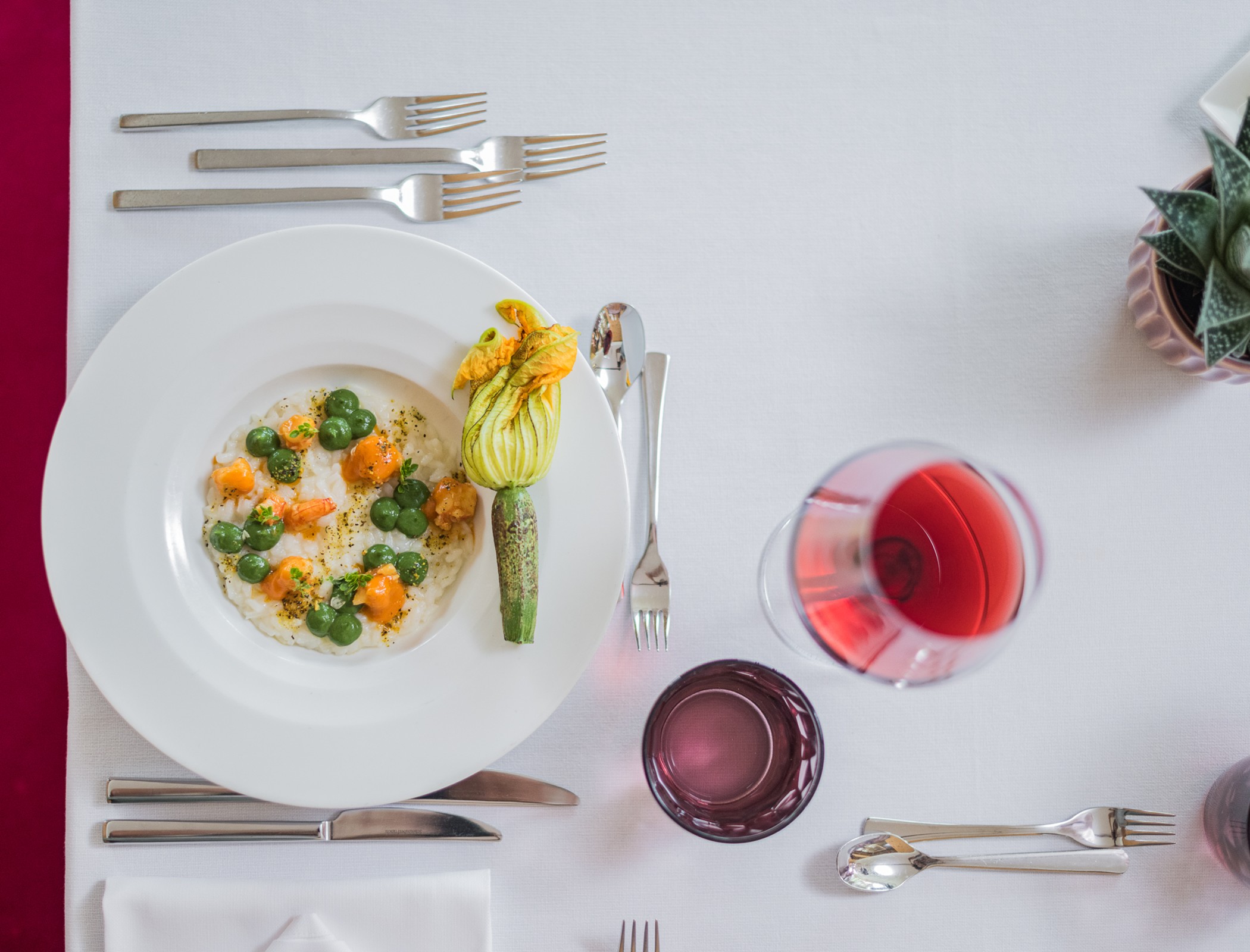 Ein weißer Teller mit einem Gourmetgericht, einem Weinglas, einem Wasserglas, Besteck und einer Topfpflanze auf einer weißen Tischdecke. - Hotel Fameli